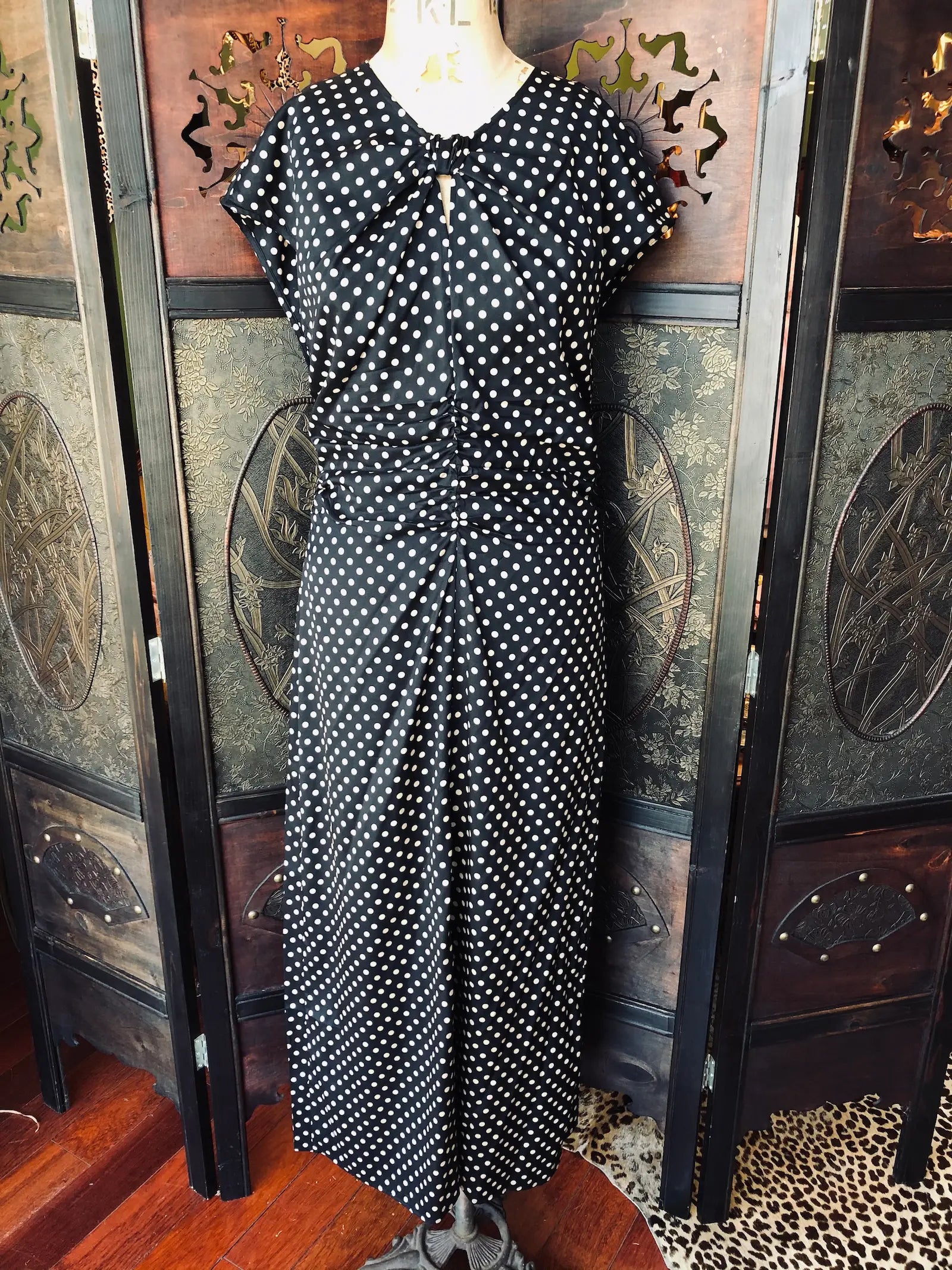 Broochella Baum und Pferdgarten silk dress size EU 38. It is black with beige polka dot print, with Ruched detailing and cap sleeves. It is pictured on a vintage mannequin. The background is an ornate, vintage, dark wood room divider. 