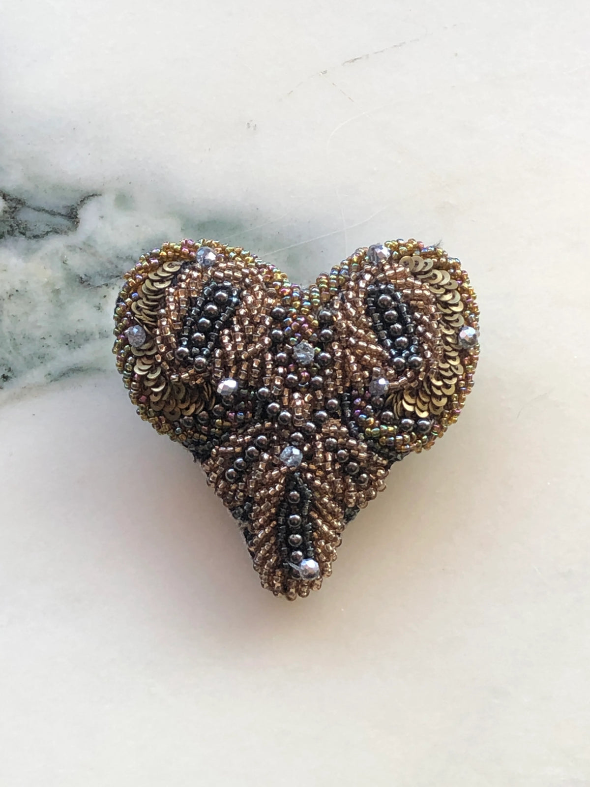 Broochella Beaded Amore Heart Brooch hand embellished with bronze, gold and gunmetal beads, sequins and crystals, photographed on a white marble background with soft grey veining.