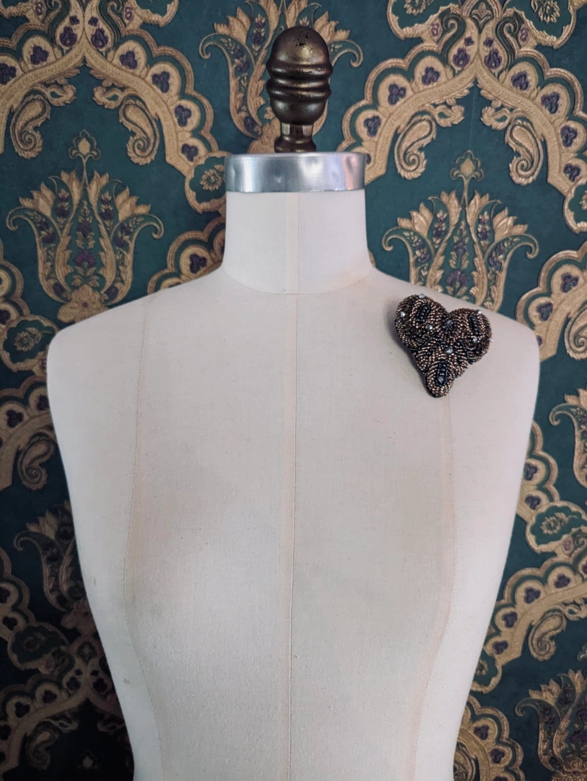 A Broochellaheart-shaped hand beaded beaded amore brooch embellished with bronze and silver beads and sequins. It is displayed on the left shoulder of a cream-colored fabric dress form against a dark teal and gold damask patterned wallpaper background.