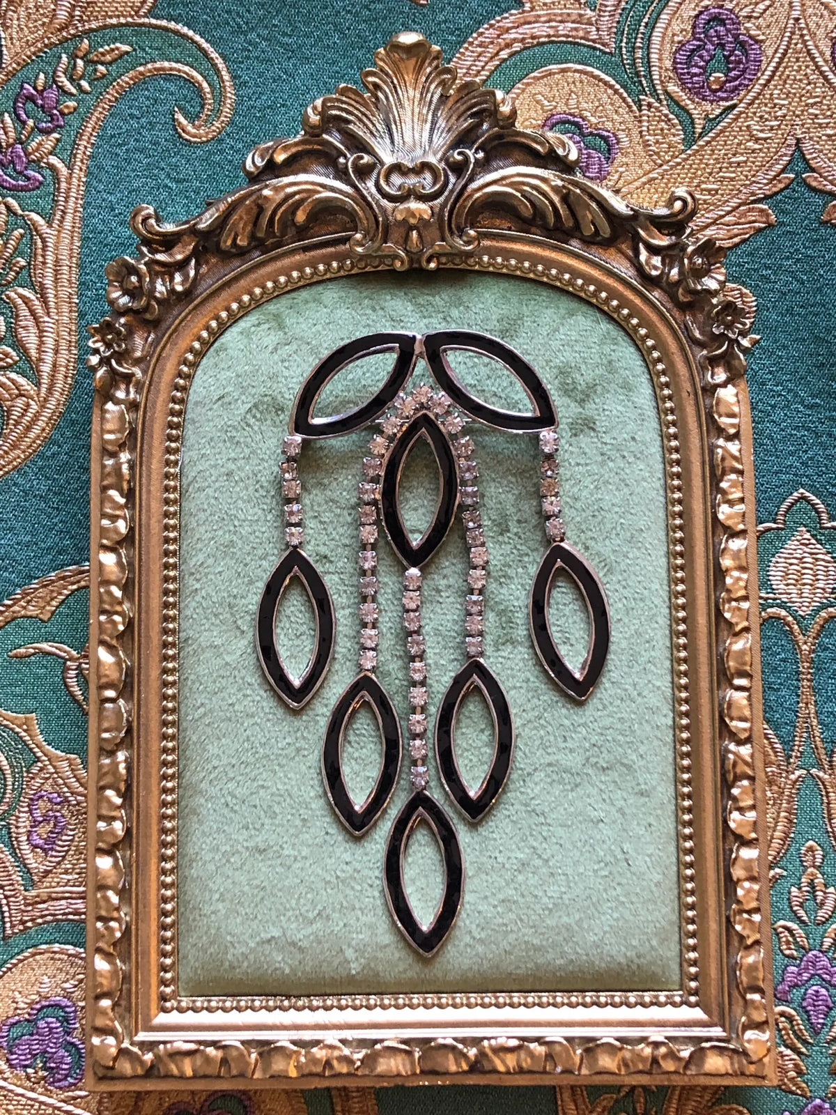 Broochella Beatrice Vintage Black Enamel Cascade Brooch featuring geometric black enamel shapes and cascading clear crystal strands in a silver-tone setting. Presented on a green velvet tray with an ornate gold frame. The background is green and gold damask wallpaper.