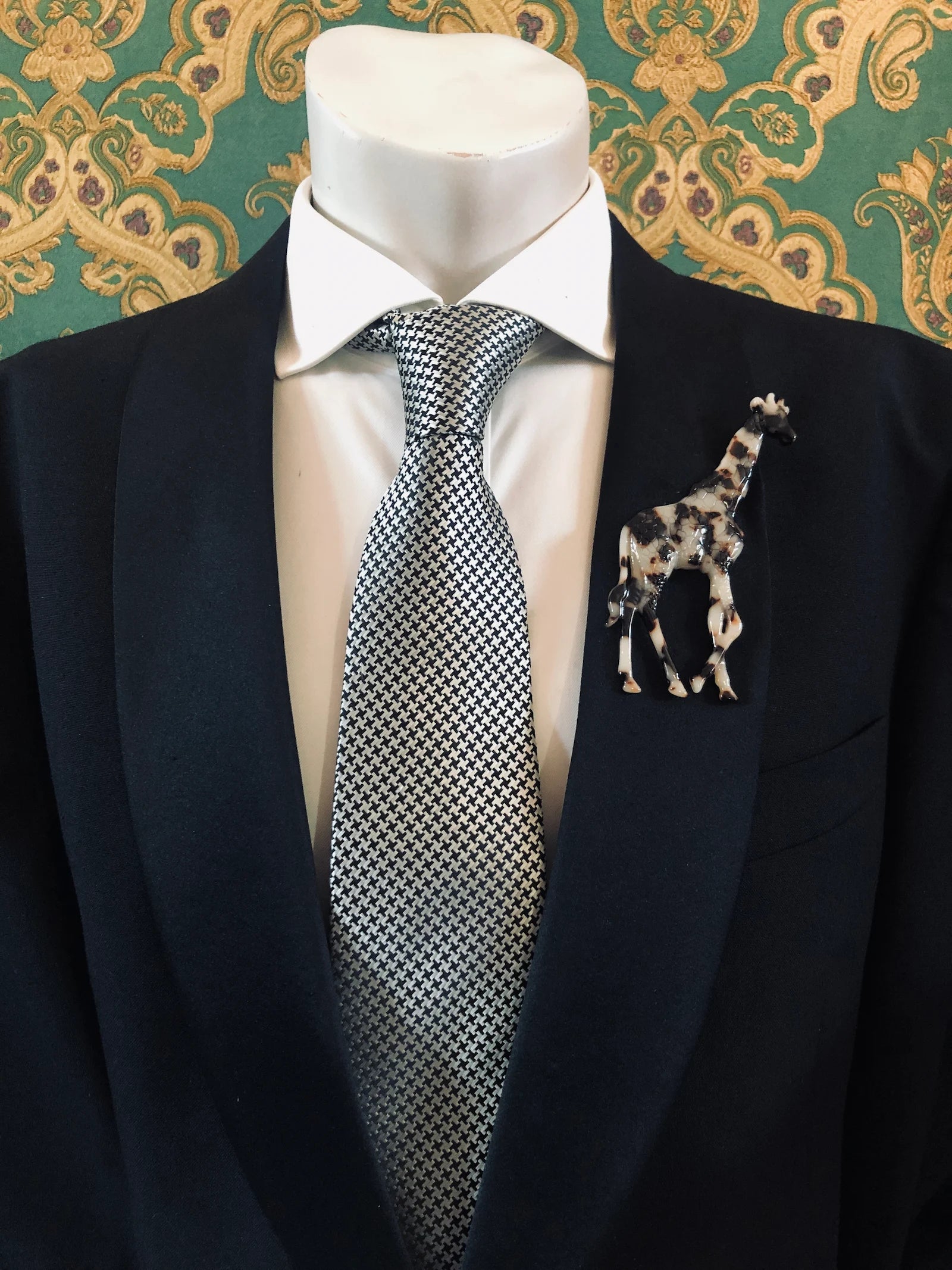 Broochella Men's beige Animalia tortoiseshell giraffe shaped brooch. It is made from Premium Italian cellulose tortoiseshell acetate. It is presented on a men's mannequin that is dressed in a black suit. The background is green and gold damask wallpaper. 