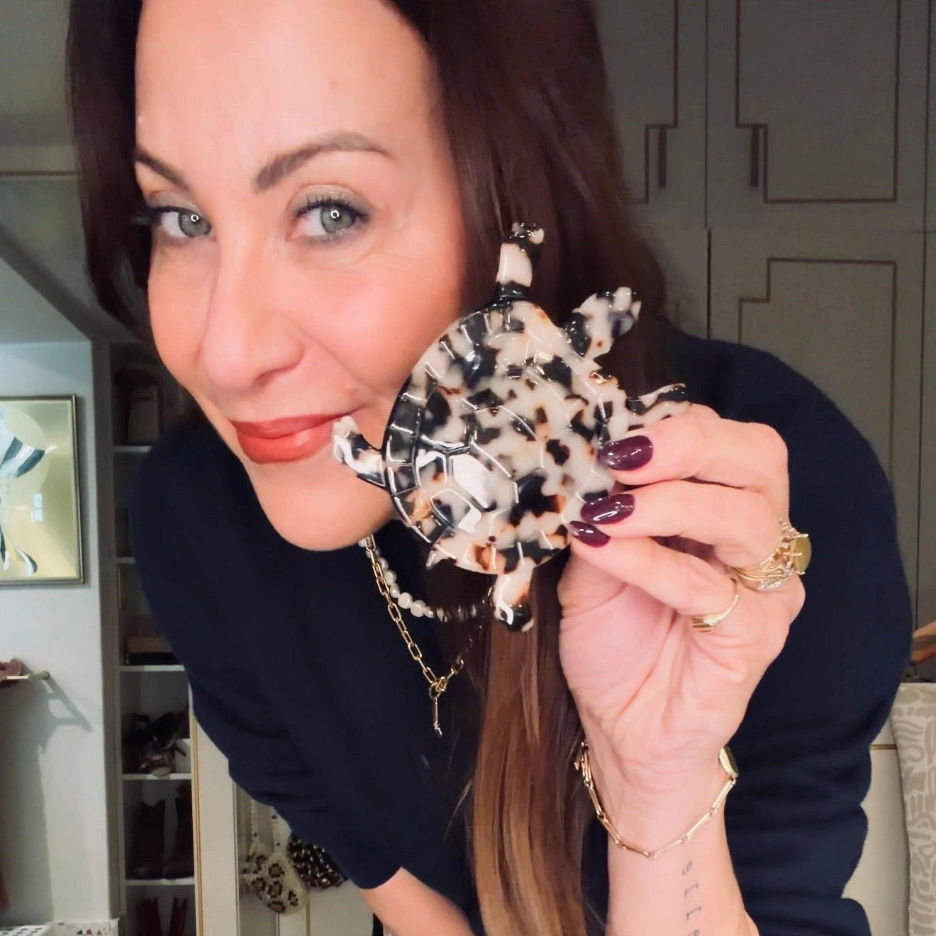 A woman holding a Broochella Animalia tortoiseshell tortoise brooch next to her face, wearing a navy blue top and gold jewelry in an indoor setting.
