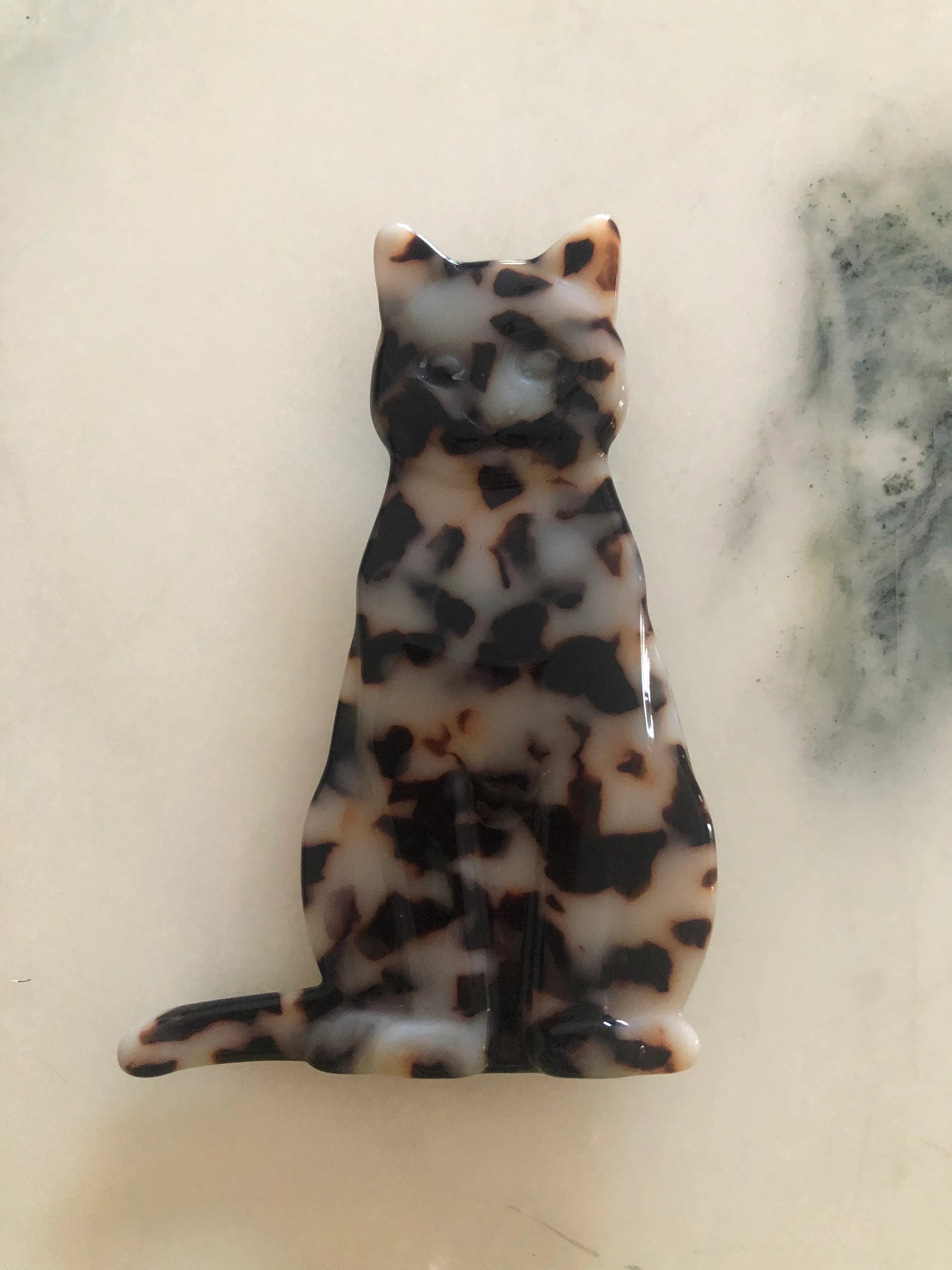 A Broochella Animalia glossy tortoiseshell cat-shaped brooch or charm with a black, brown, and cream mottled pattern, displayed on a light marble surface.