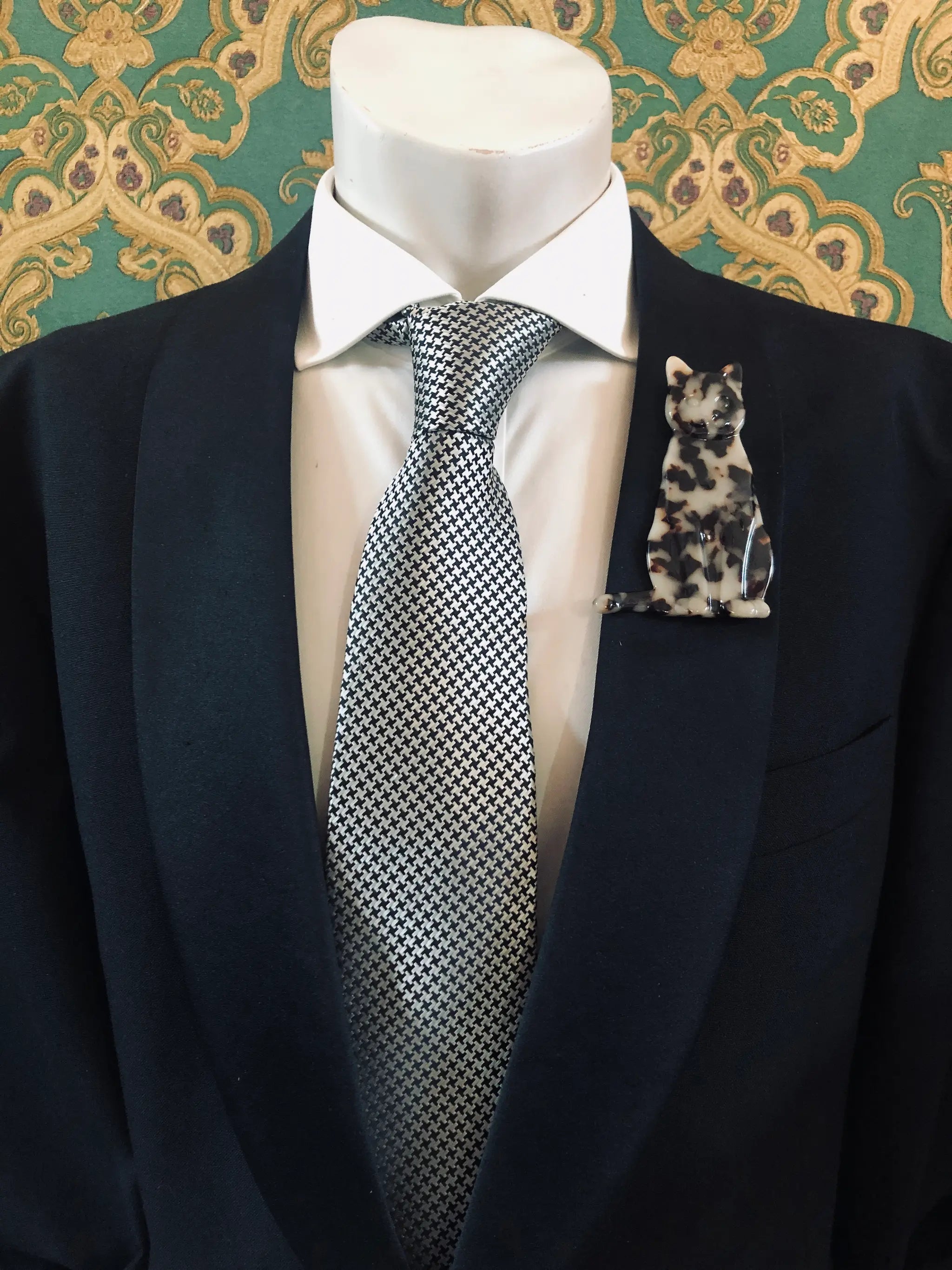 Broochella Men's beige Animalia tortoiseshell cat shaped brooch. It is made from Premium Italian cellulose tortoiseshell acetate. It is presented on a men's mannequin that is dressed in a black suit. The background is green and gold damask wallpaper. 