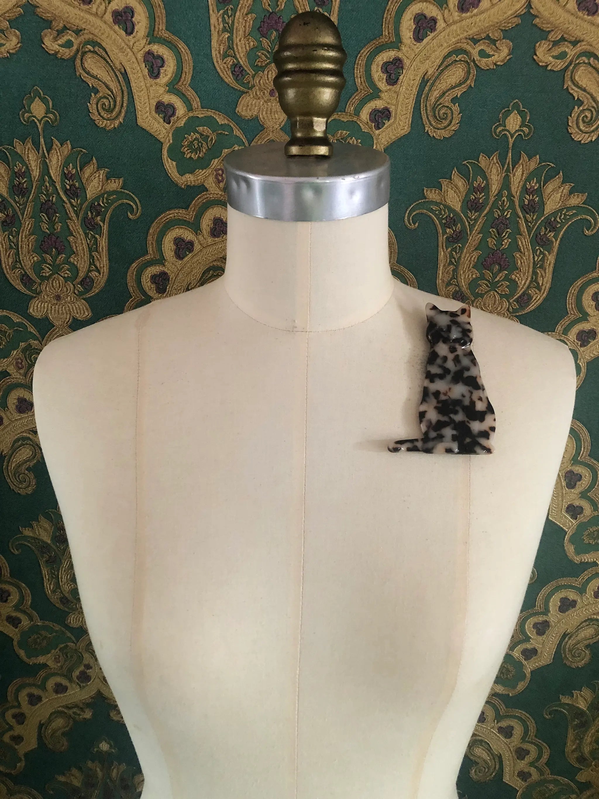 A Broochella Animalia black, brown and beige tortoiseshell cat-shaped brooch made from premium Italian cellulose acetate. It  displayed on a mannequin. The background is green and gold damask wallpaper. 