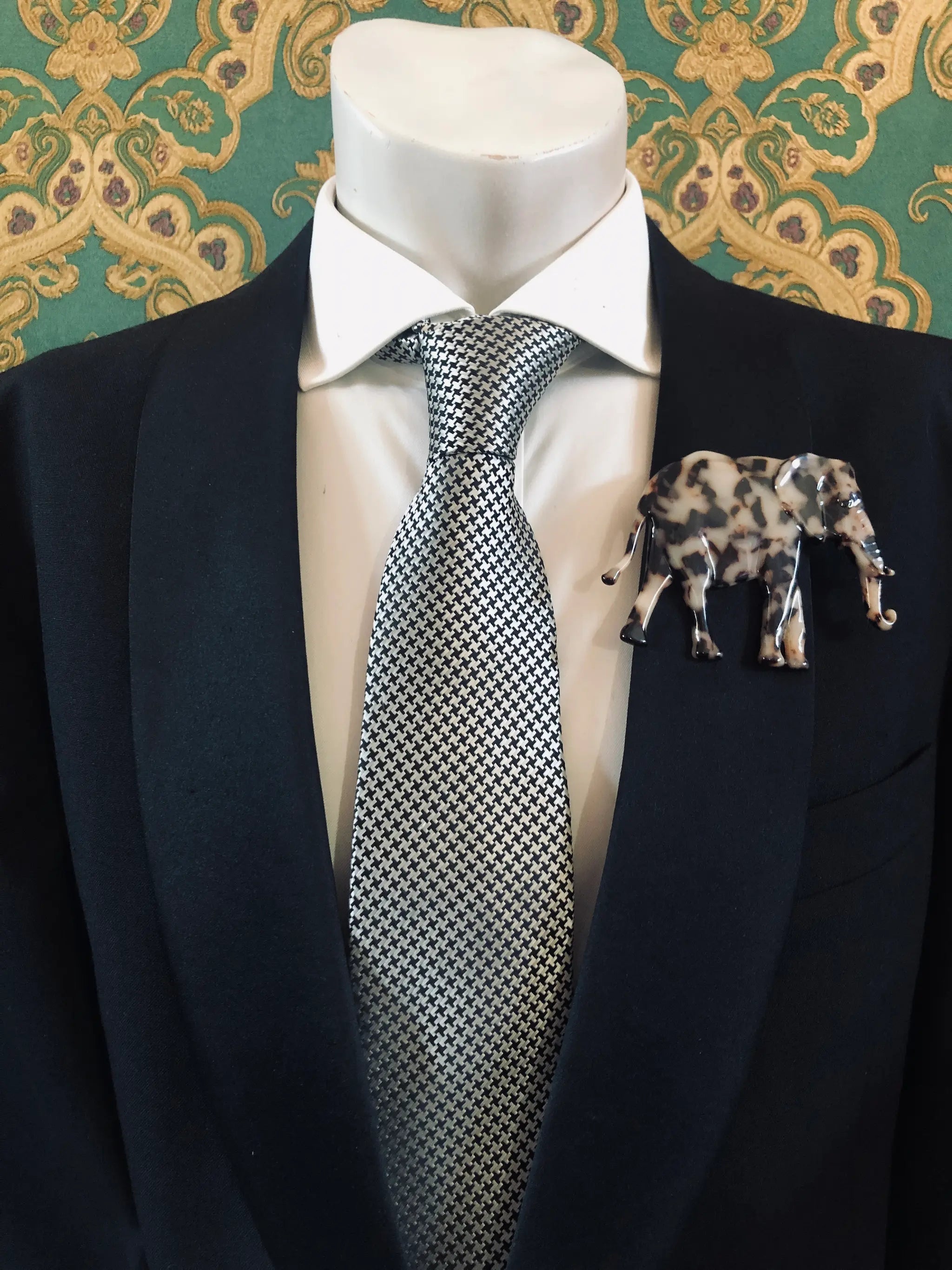 Broochella Men's beige Animalia tortoiseshell elephant shaped brooch. It is made from Premium Italian cellulose tortoiseshell acetate. It is presented on a men's mannequin that is dressed in a black suit. The background is green and gold damask wallpaper. 