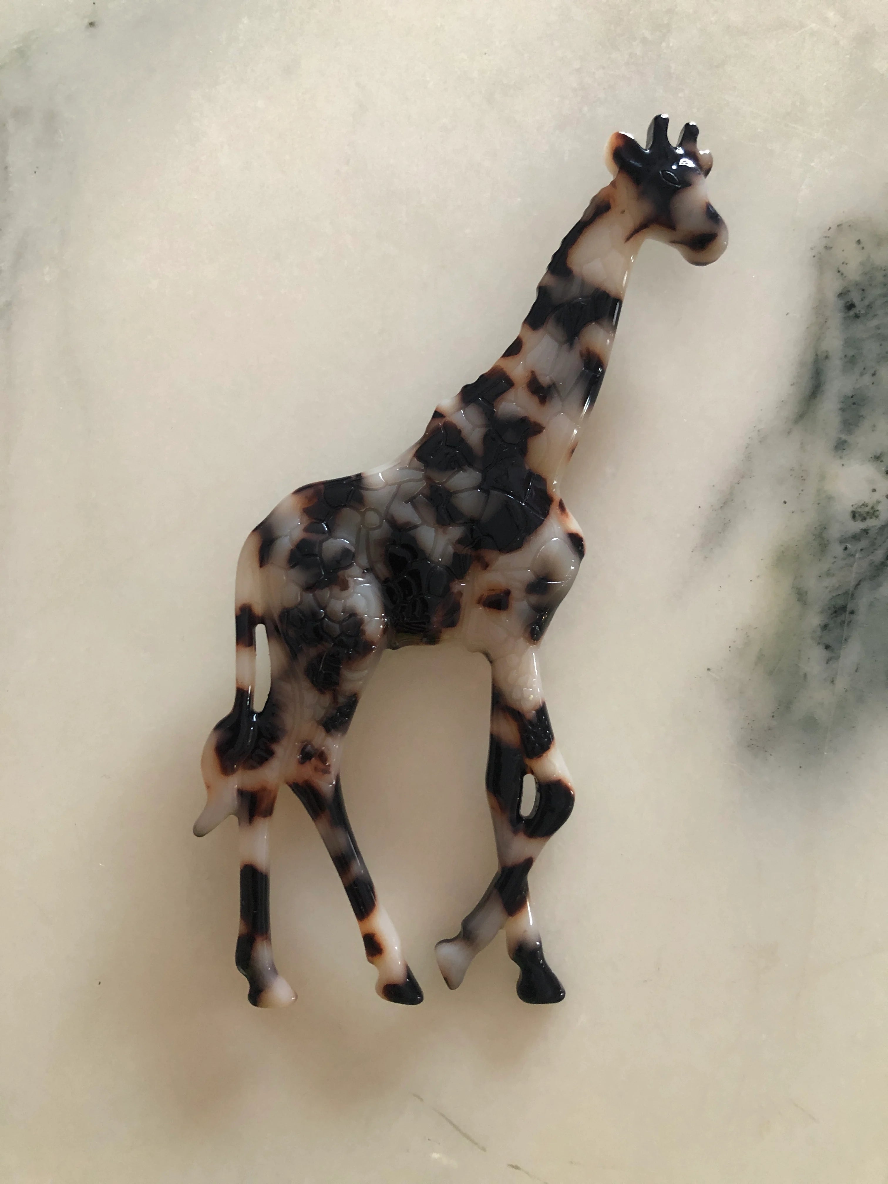 Broochella Men's beige Animalia tortoiseshell giraffe shaped brooch. It is made from Premium Italian cellulose tortoiseshell acetate. It is presented on a white marble surface. 