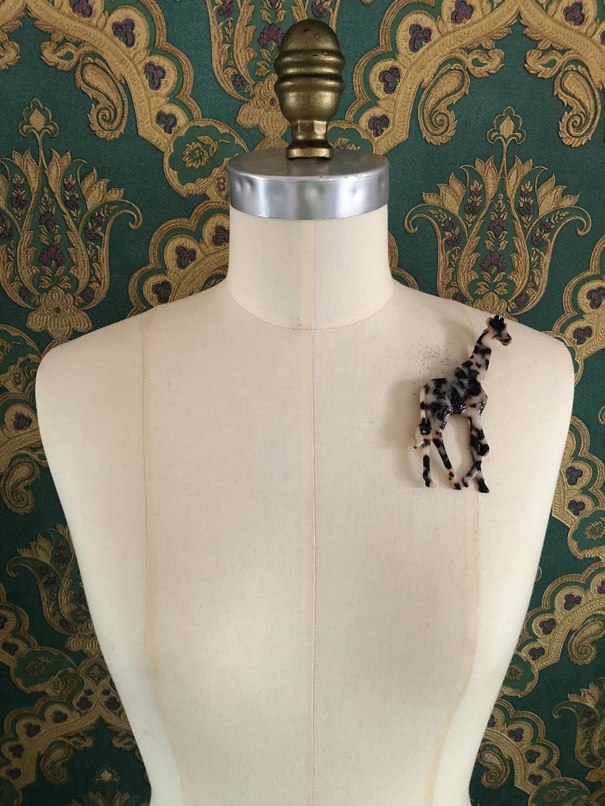 A Broochella Animalia Collection standing giraffe brooch in brown, black and beige tortoiseshell Italian cellulose acetate with sculpted spot patterning measuring 12cm x 7cm. It is pinned on a mannequin. The background is green and gold damask wallpaper. 