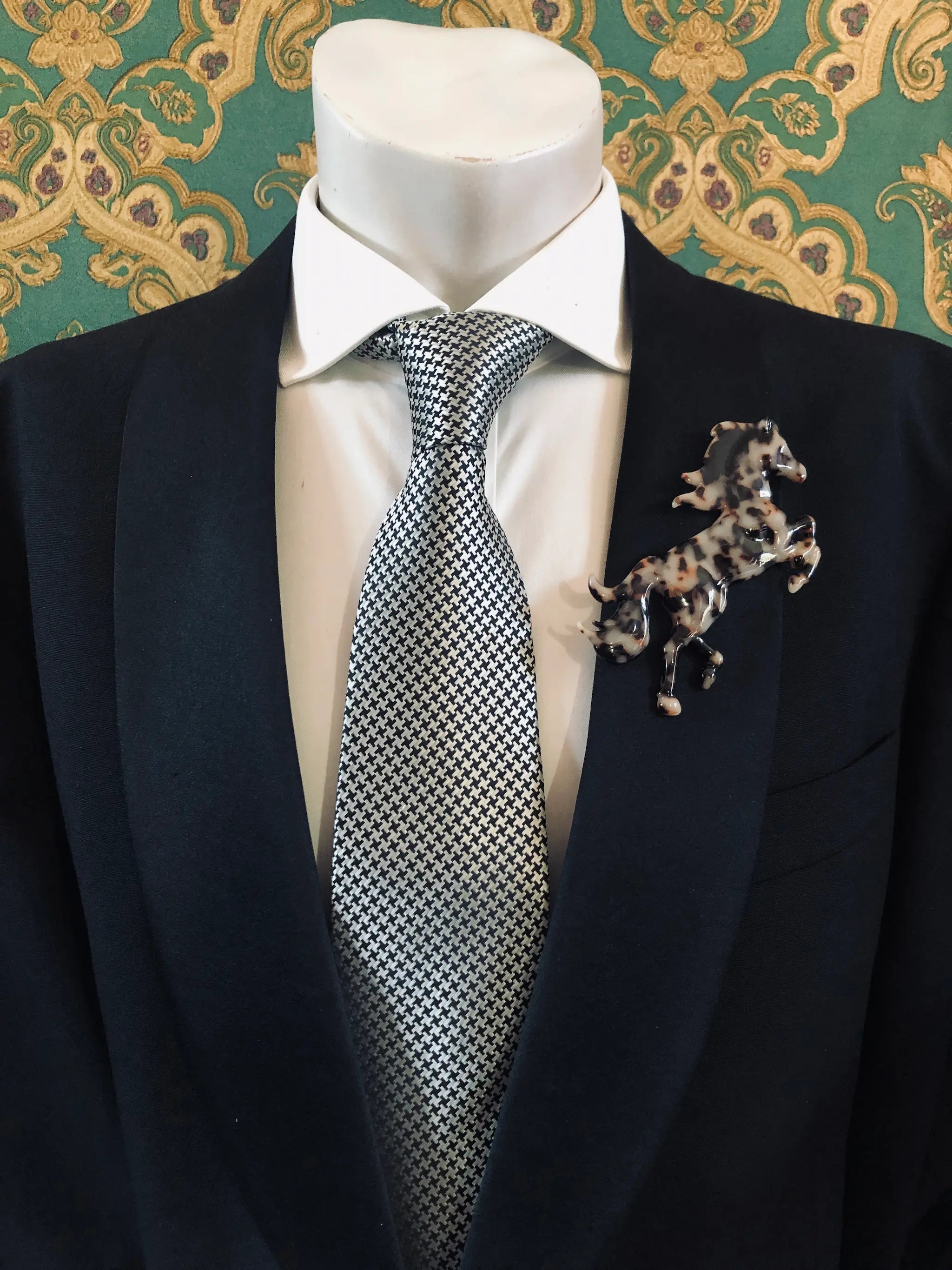 Broochella Men's Animalia beige,  brown and black tortoiseshell figural horse brooch. It is pinned to a men's mannequin that is dressed in a black suit. The background is green and gold damask wallpaper. 