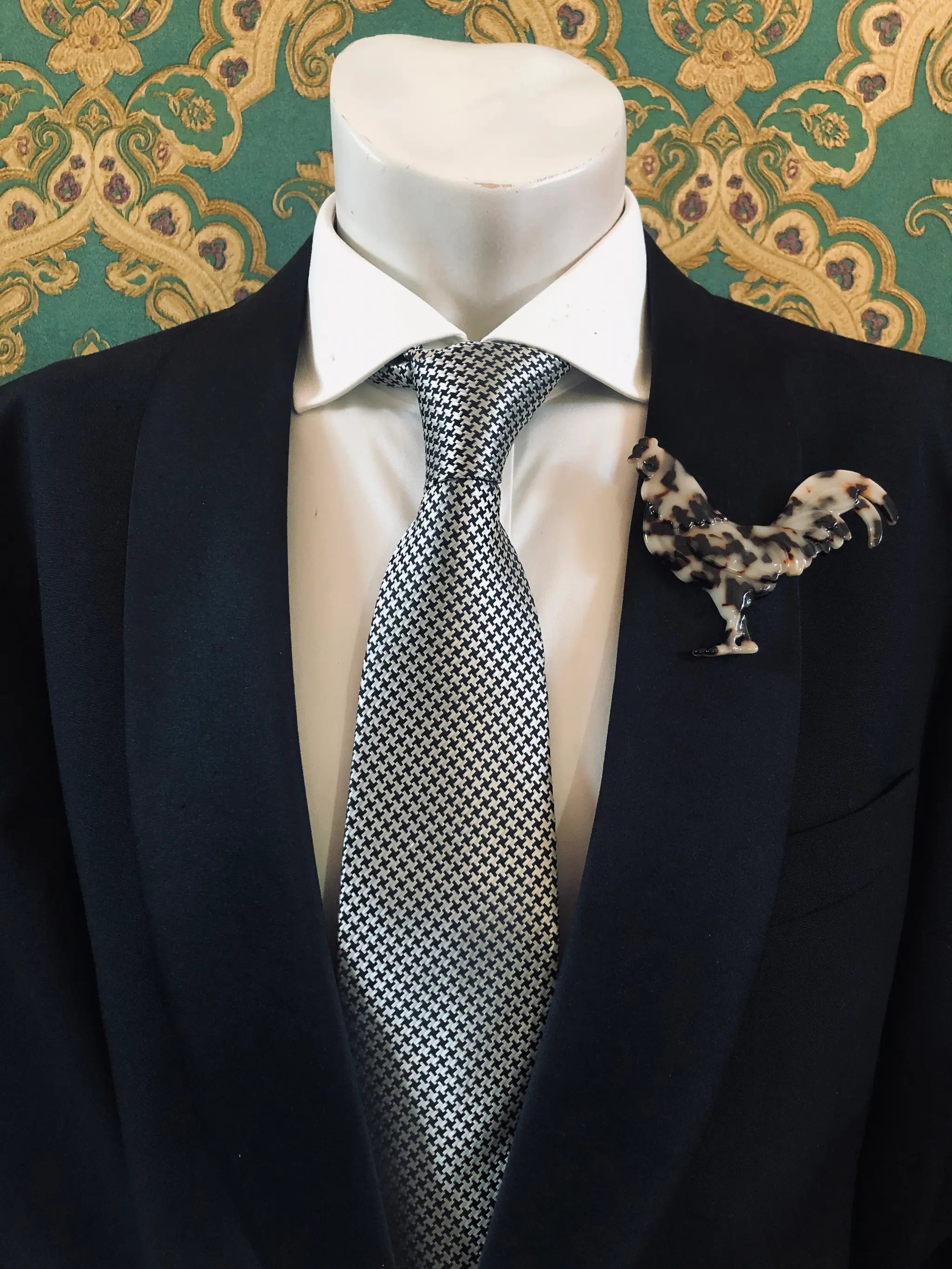 A Broochella Animalia Men's tortoiseshell rooster brooch displayed on a white mannequin torso, featuring a black  suit jacket, a white dress shirt, a silver and black checkered tie against a green and gold patterned wallpaper background.