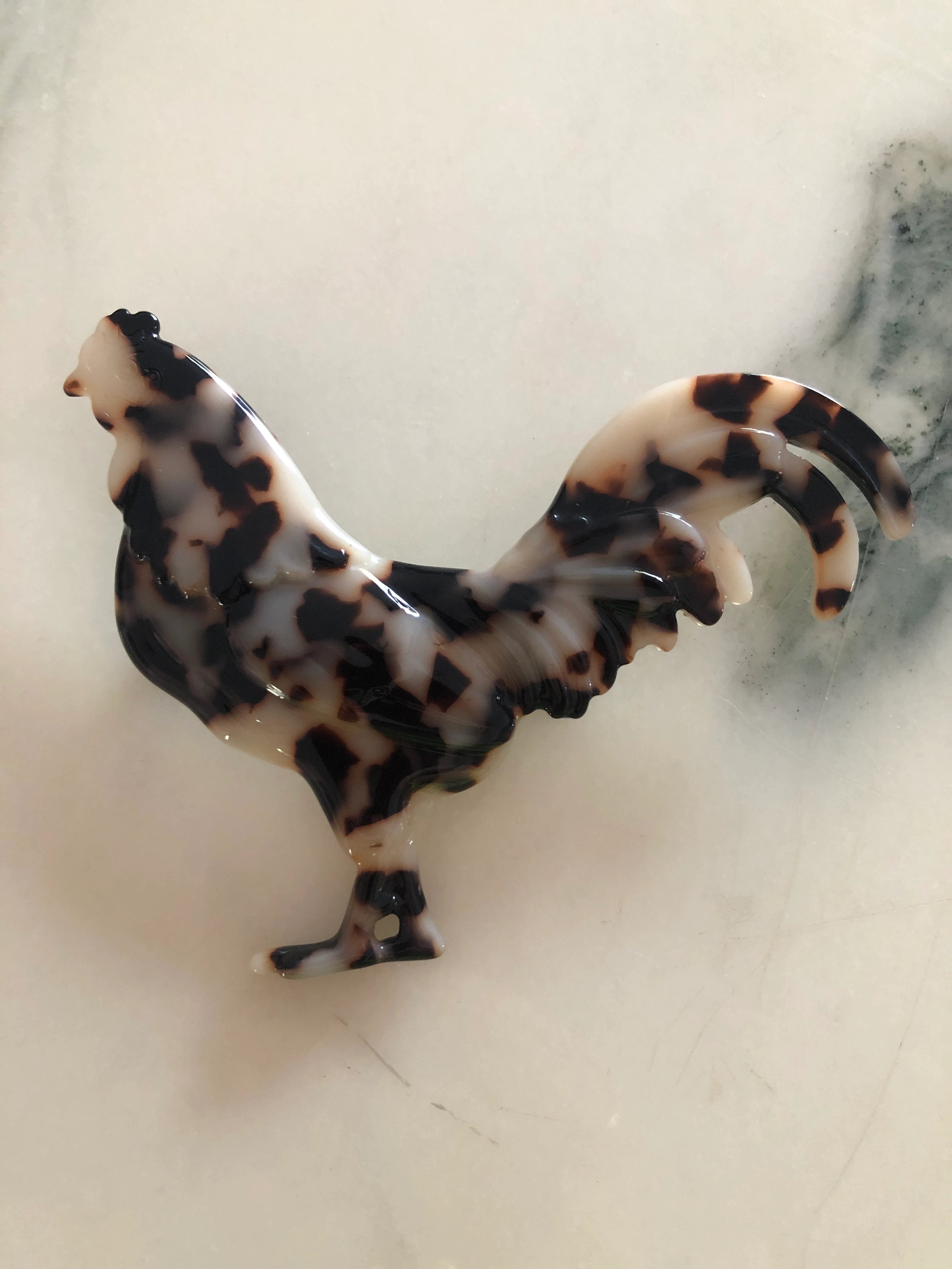 A Broochella Animalia Men's tortoiseshell, figural  rooster brooch, made from premium Italian cellulose acetate. It is displayed on a white marble table