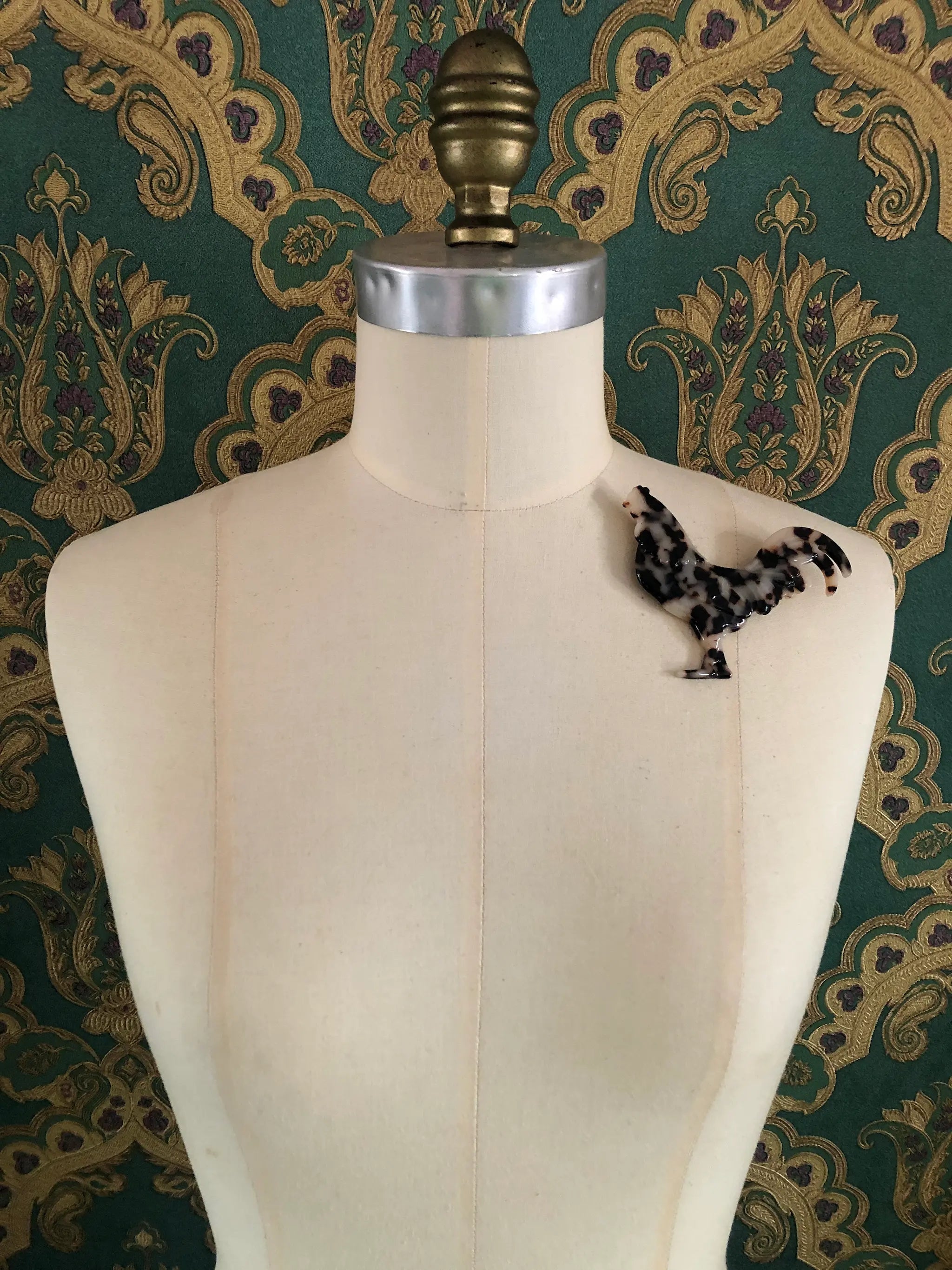 Broochella Animalia handmade, tortoiseshell, Italian cellulose acetate rooster brooch in beige. It is pinned to a mannequin. The background is green and gold damask wallpaper.