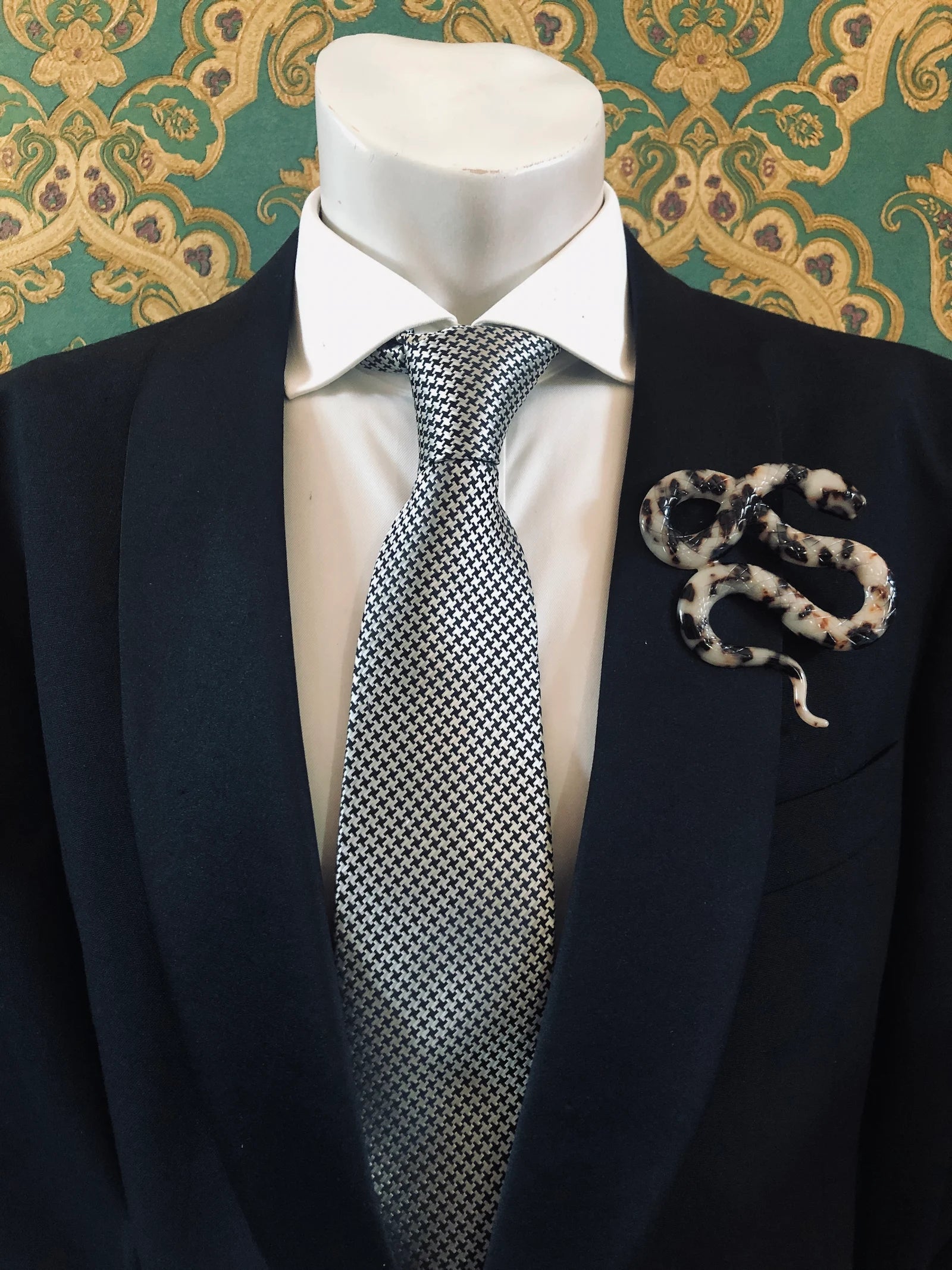 Broochella Men's beige Animalia tortoiseshell snake shaped brooch. It is made from Premium Italian cellulose tortoiseshell acetate. It is presented on a men's mannequin that is dressed in a black suit. The background is green and gold damask wallpaper. 