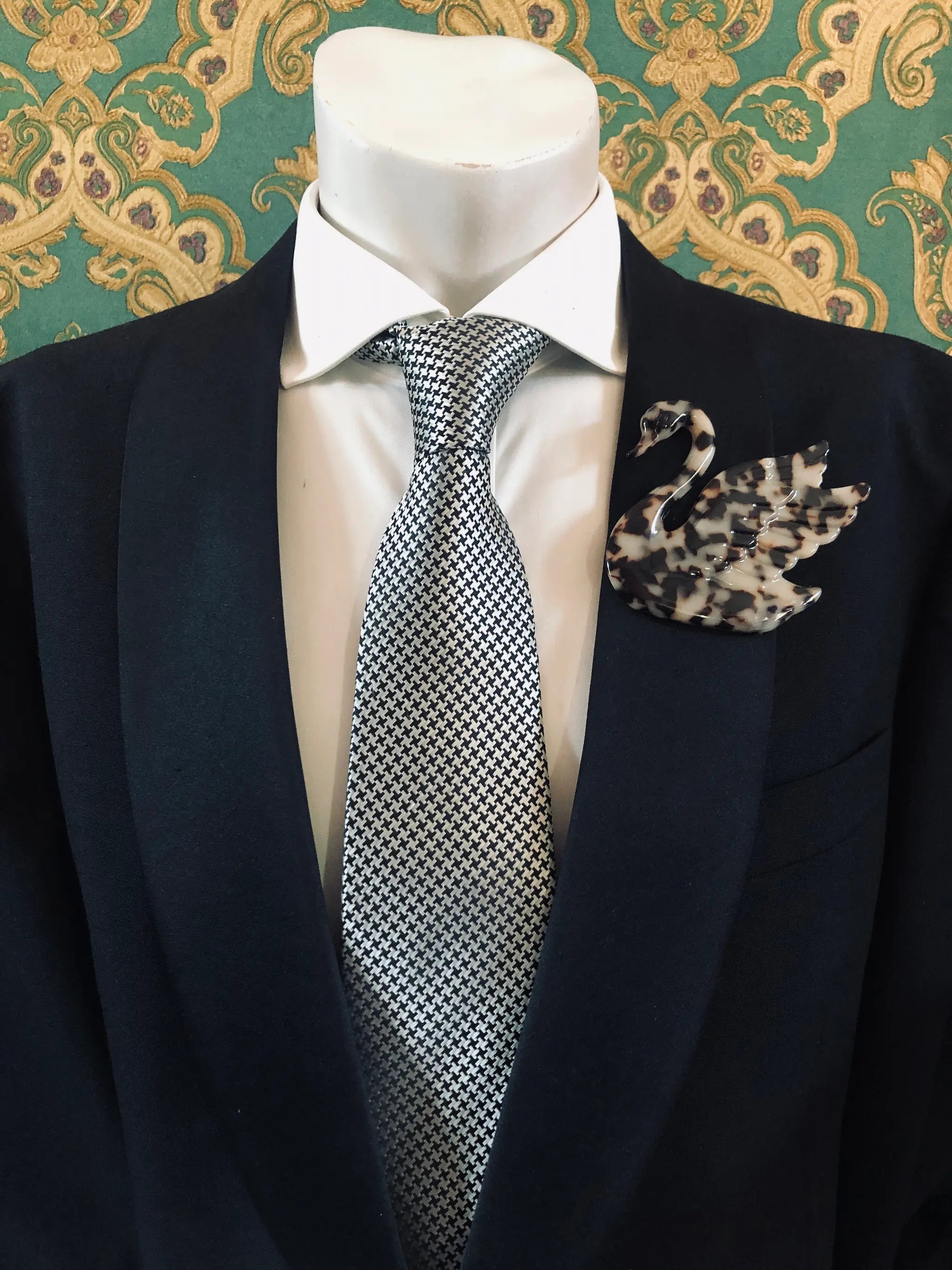 A Broochella Animalia Men's tortoiseshell beige swan brooch displayed on a white mannequin torso, featuring a black  suit jacket, a white dress shirt, a silver and black checkered tie against a green and gold patterned wallpaper background.