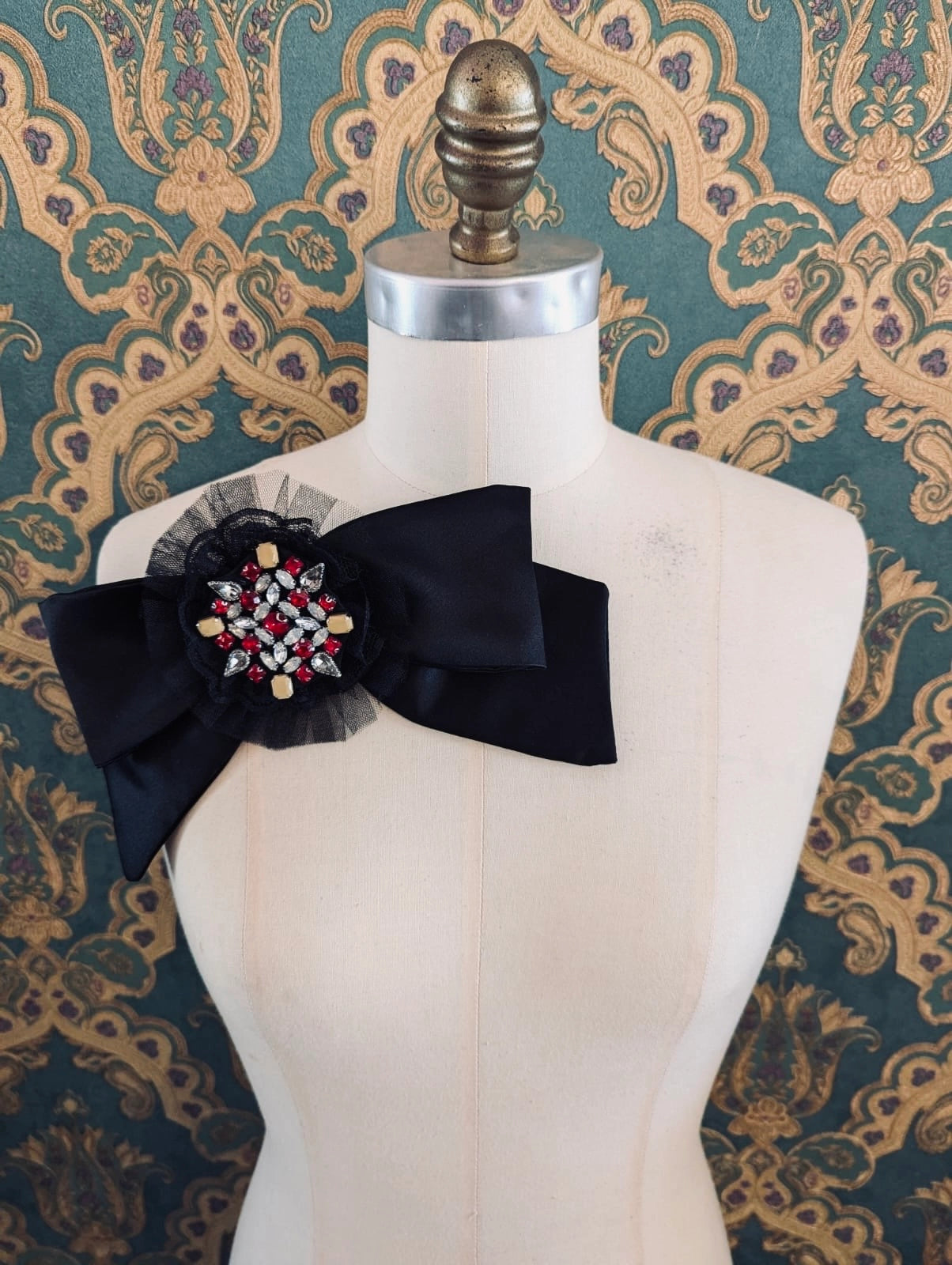 Broochella viva loren bejewelled black bow brooch. It has a red a crystal embellished brooch at the centre of the bow. It is pinned to a mannequin. The background is green and gold damask wallpaper.