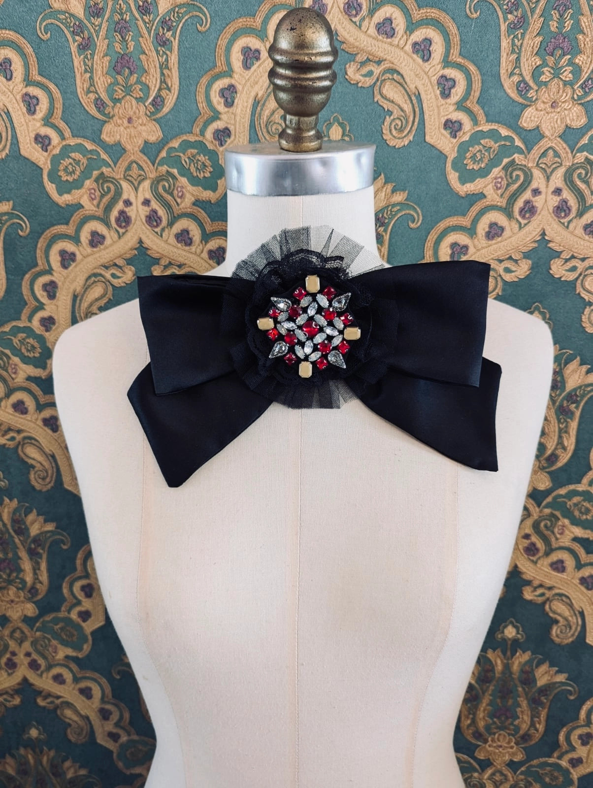 Broochella viva loren bejewelled black bow brooch. It has a red a crystal embellished brooch at the centre of the bow. It is pinned to a mannequin. The background is green and gold damask wallpaper.