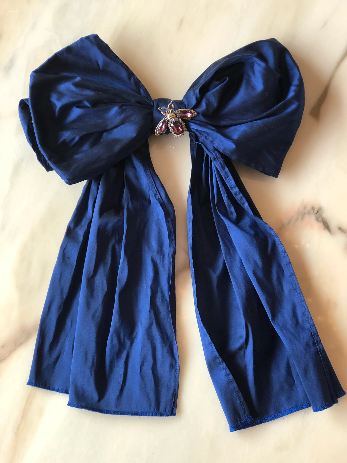 Broochella Bejewelled Fashion Bow Brooch Pin in rich blue with an ornate jewelled centre and long flowing tails, photographed on a white marble background.