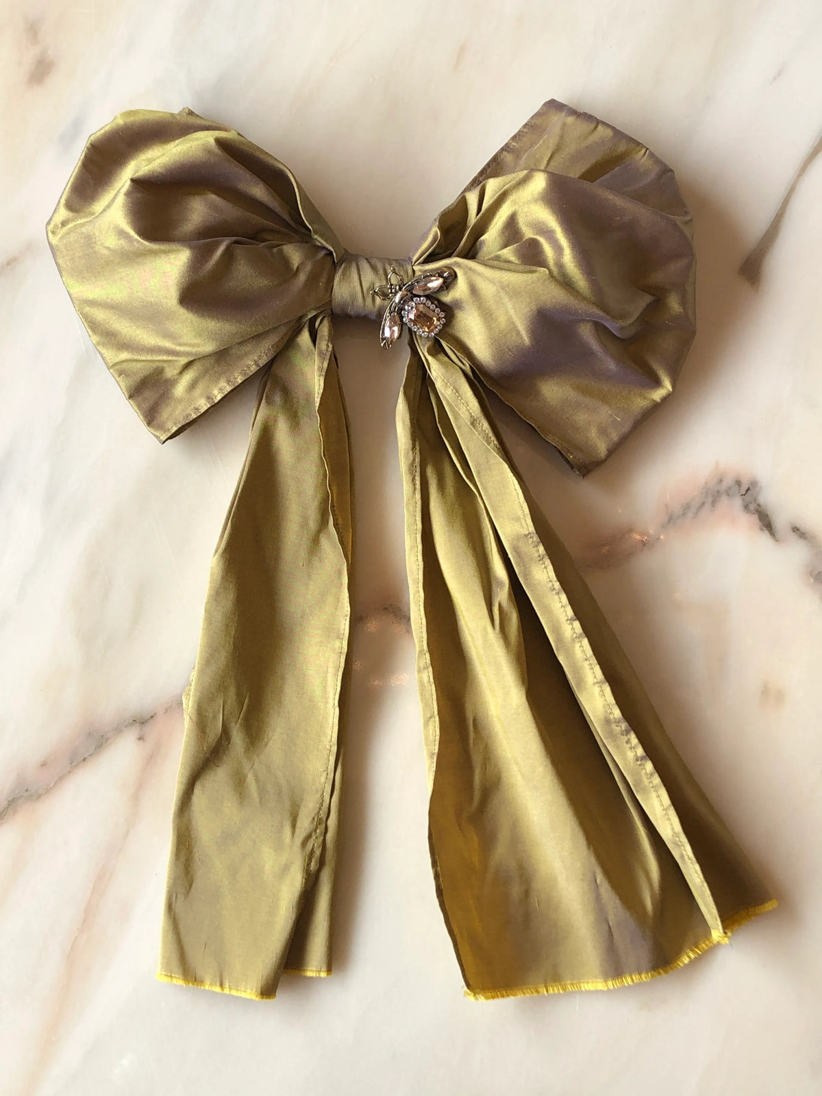 Broochella Bejewelled Fashion Bow Brooch Pin in gold with an ornate jewelled centre and long flowing tails, photographed on a white marble background.