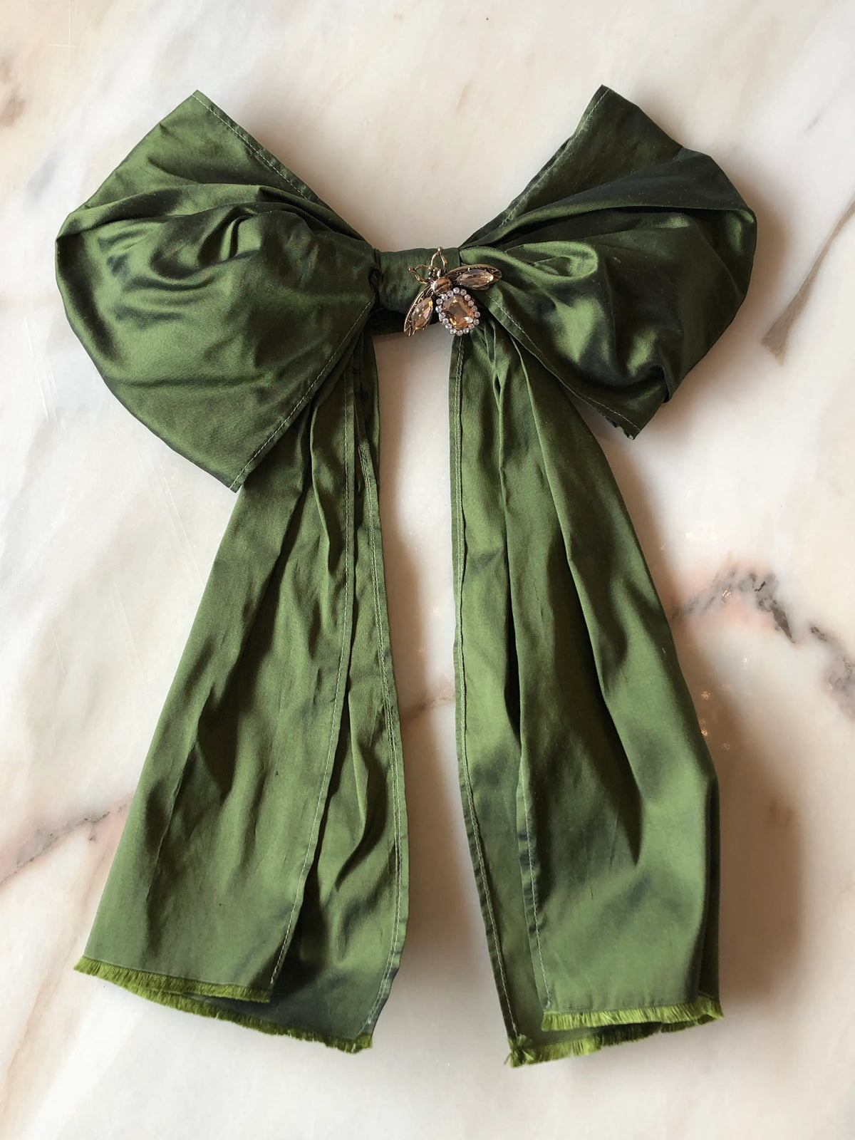 Broochella Bejewelled Fashion Bow Brooch Pin in olive with an ornate jewelled centre and long flowing tails, photographed on a white marble background.