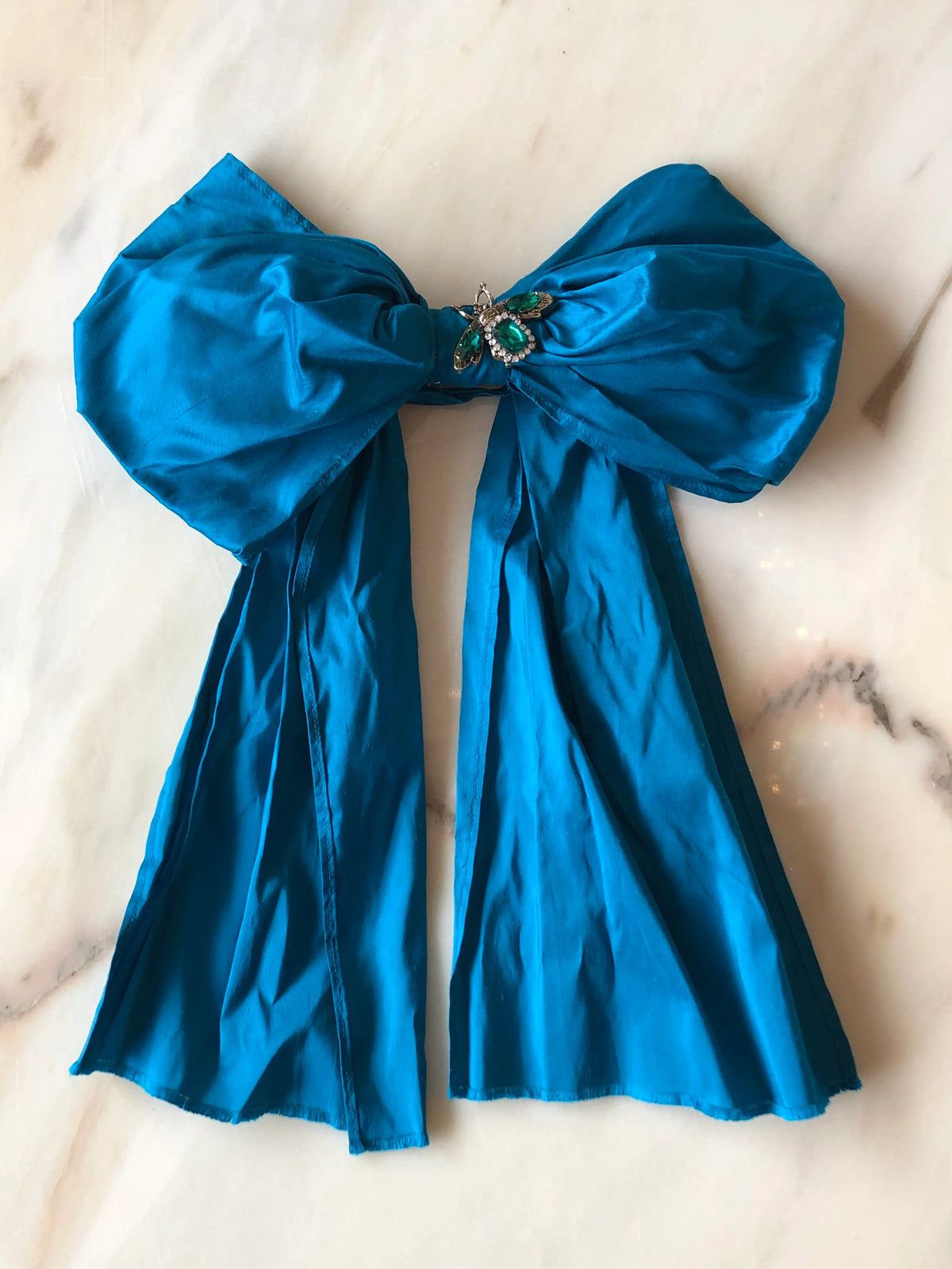 Broochella Bejewelled Fashion Bow Brooch Pin in turquoise with an ornate jewelled centre and long flowing tails, photographed on a white marble background.