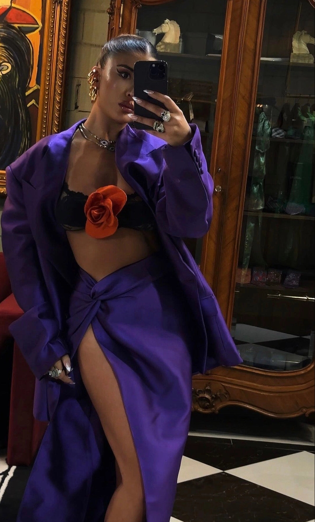 A woman takes a mirror selfie wearing a purple satin-like outfit with a Broochella  Bellucci orange, handmade, silk flower brooch on the bra top, a matching high-slit skirt, and an oversized purple blazer. The setting is an indoor room with a wooden display cabinet and checkered floor.