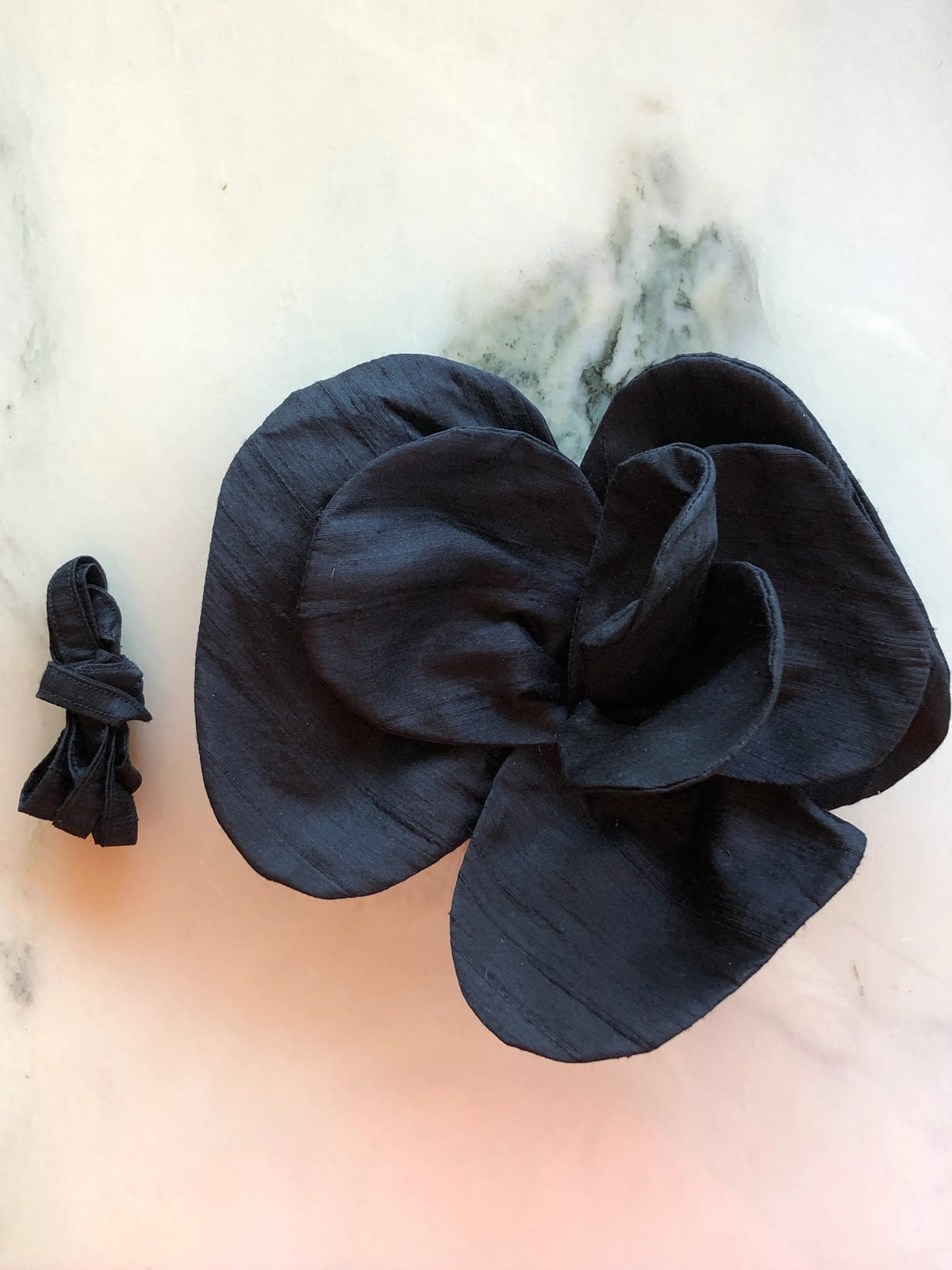 Small black Broochella Bellucci Silk-Flower Choker & Brooch with sculptural silk petals and matching silk choker ribbon, photographed on a white marble background with soft grey veining.