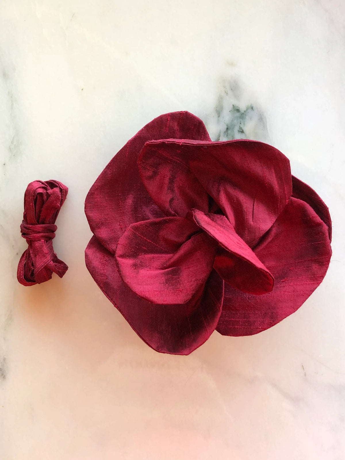 Small burgundy Broochella Bellucci Silk-Flower Choker & Brooch with sculptural silk petals and matching silk choker ribbon, photographed on a white marble background with soft grey veining.