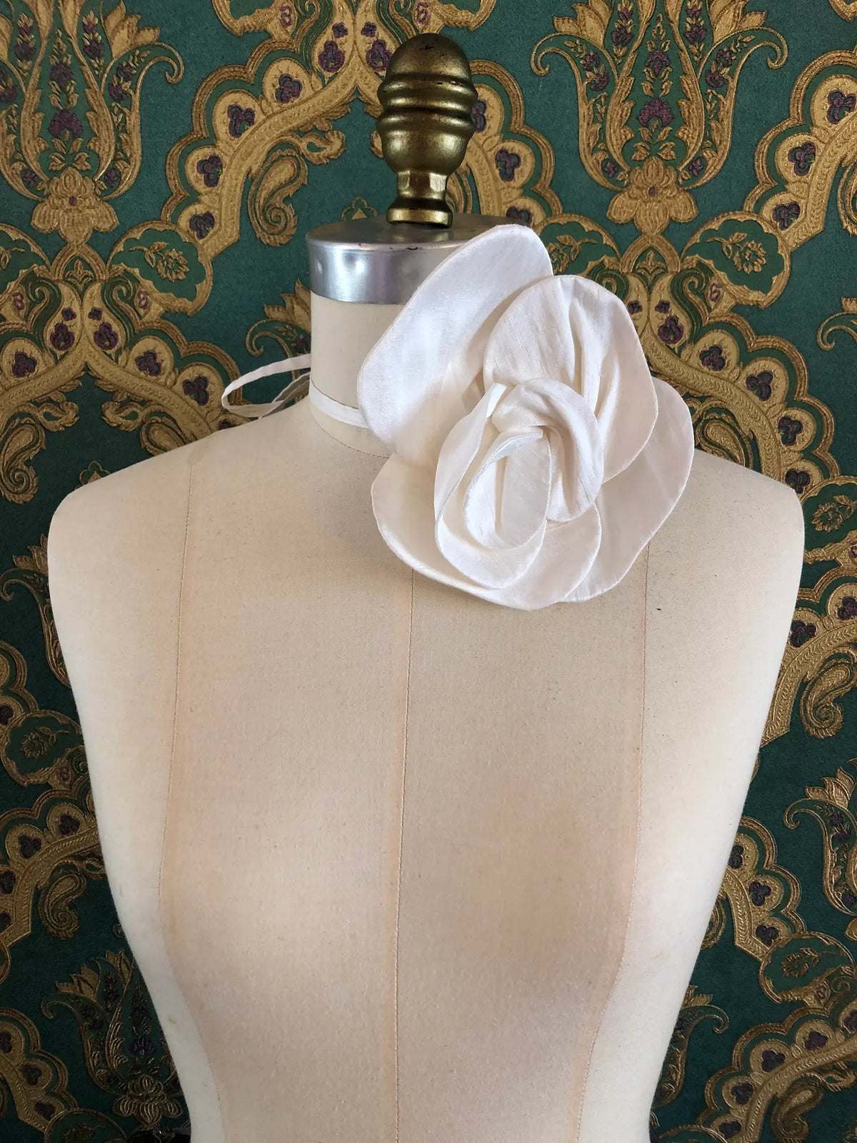 Small ivory Broochella Bellucci Silk-Flower Choker & Brooch with sculptural silk petals and matching silk choker ribbon, photographed on a mannequin. The background is green and gold damask wallpaper.
