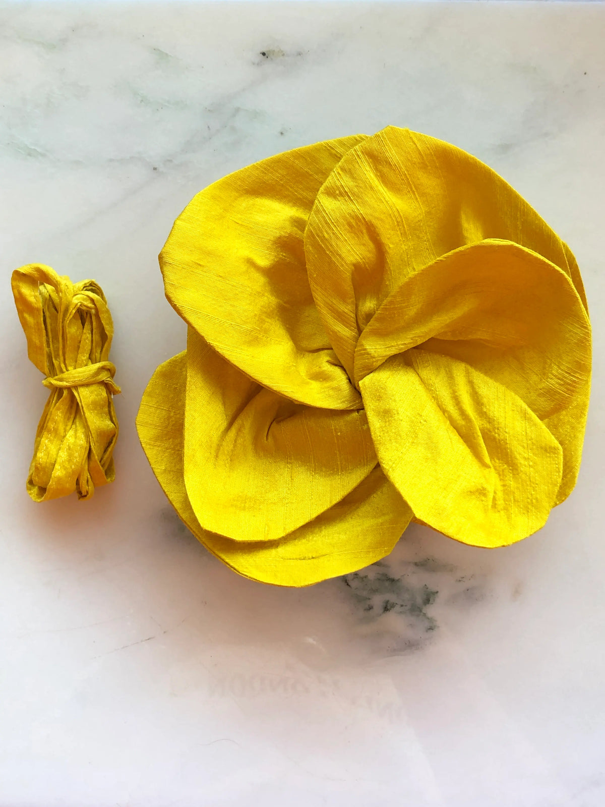 Small yellow Broochella Bellucci Silk-Flower Choker & Brooch with sculptural silk petals and matching silk choker ribbon, photographed on a white marble background with soft grey veining.
