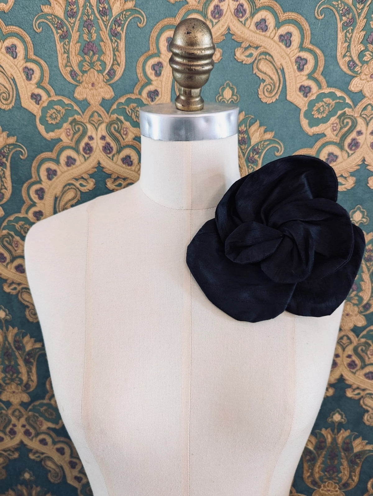 Broochella Bellucci small black silk flower brooch with sculptural layered petals, styled on a cream mannequin against an ornate teal wallpaper background.