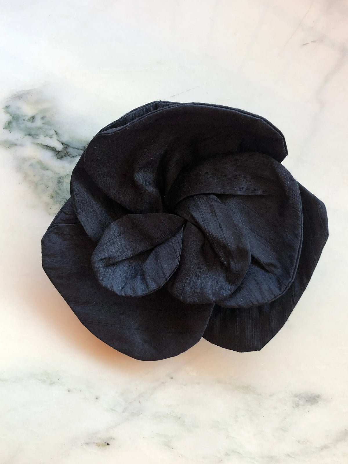 Broochella Bellucci small black silk flower brooch with oversized sculptural petals and subtle silk sheen, photographed on a white marble background with grey veining.
