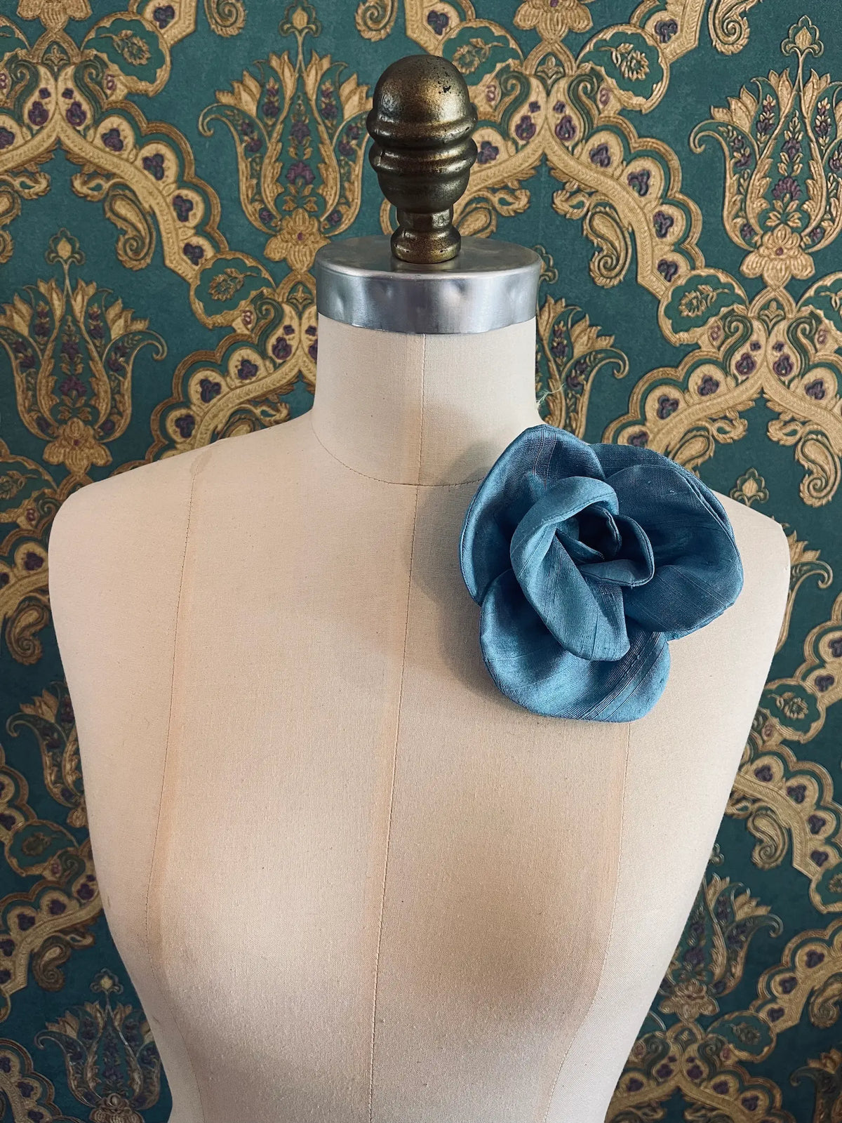 Broochella Bellucci small blue silk flower brooch with softly layered petals, styled on a cream mannequin against an ornate teal wallpaper background.