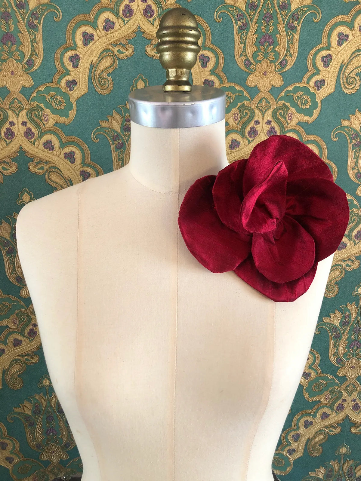 Broochella Bellucci small burgundy silk flower brooch with rich sculptural petals, styled on the shoulder of a cream mannequin against ornate teal wallpaper.