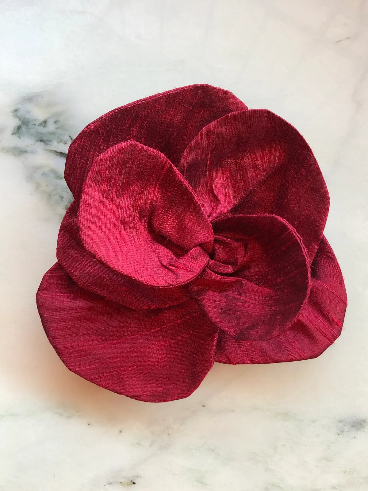 Broochella Bellucci small burgundy silk flower brooch with deep red sculptural petals and lustrous finish, photographed on a white marble background with grey veining.