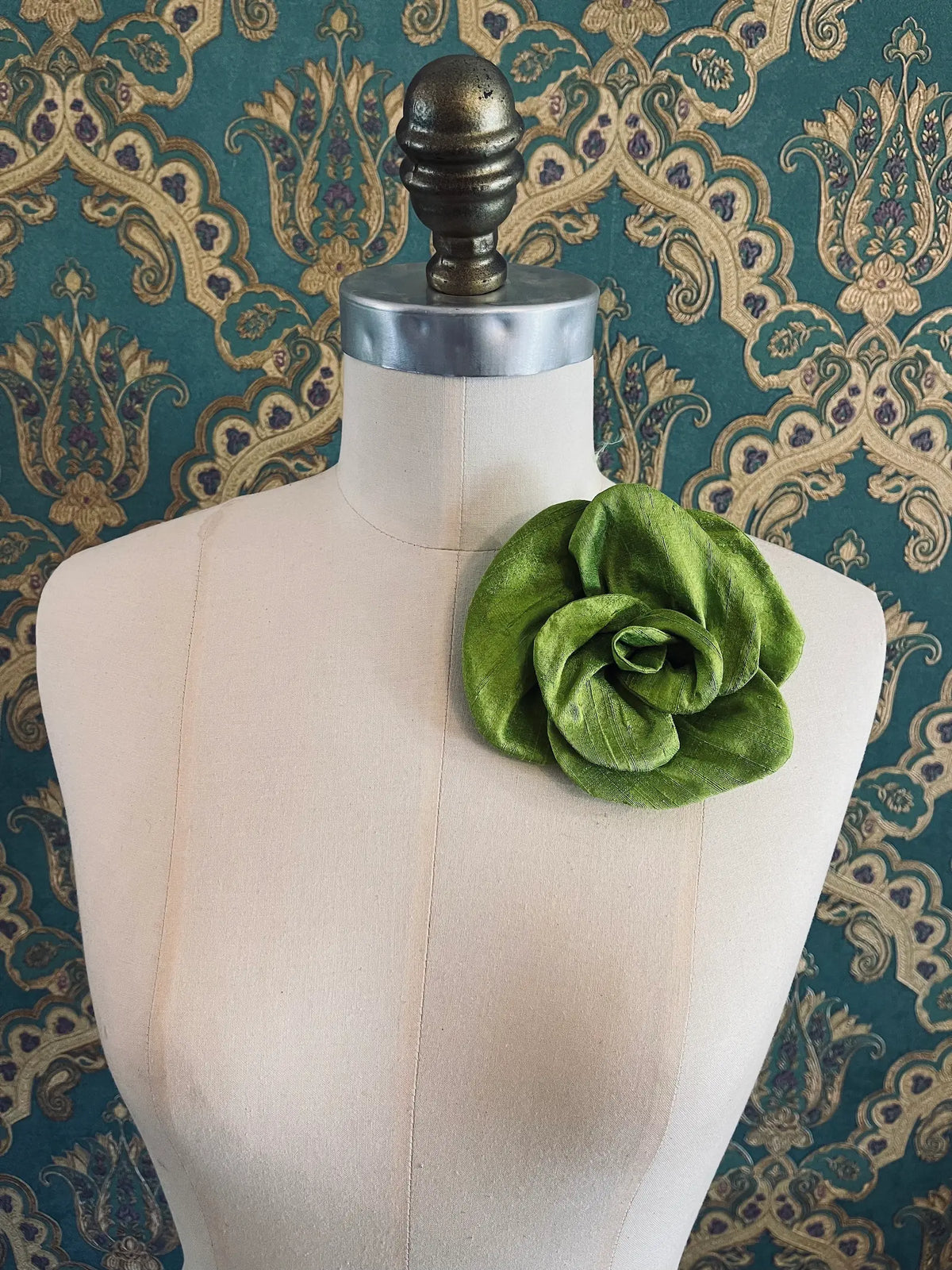 Broochella Bellucci small chartreuse silk flower brooch with lustrous layered petals, styled on a cream mannequin against an ornate teal wallpaper background.