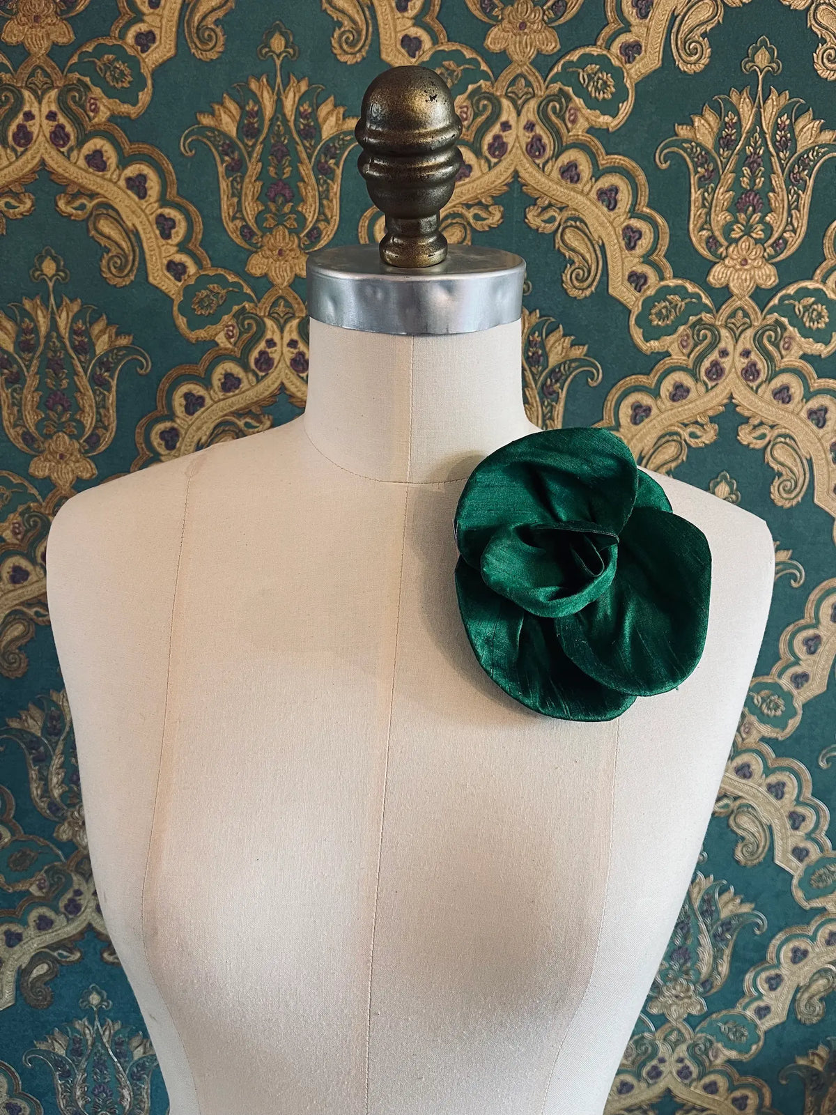 Broochella Bellucci small emerald green silk flower brooch with sculptural petals, styled on the shoulder of a cream mannequin against ornate teal wallpaper.