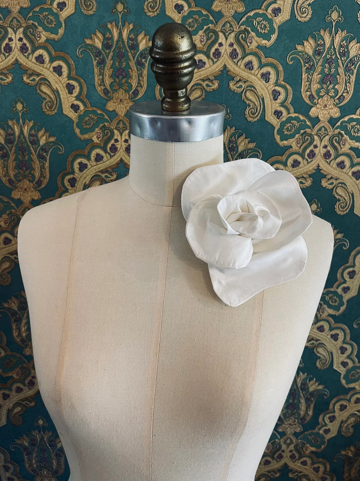 Broochella Bellucci small yellow silk flower brooch with lustrous sculptural petals, styled on the shoulder of a cream mannequin against ornate teal wallpaper.