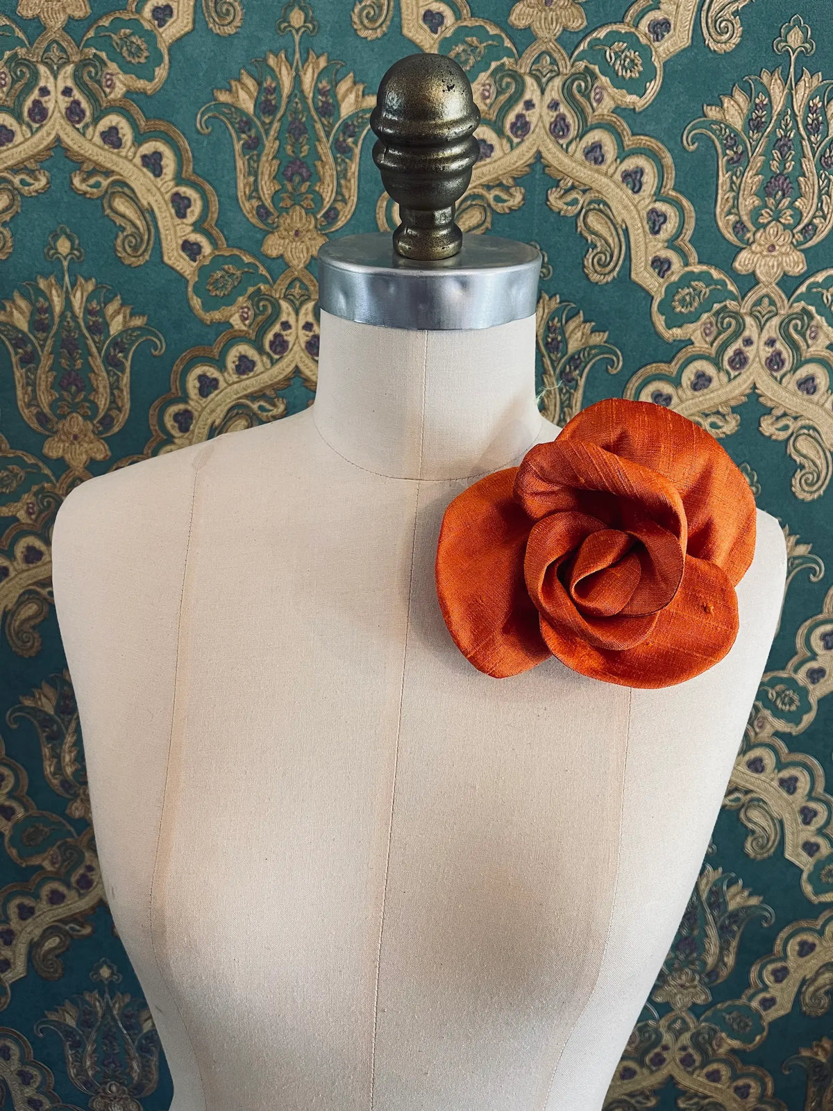 Broochella Bellucci small orange silk flower brooch with lustrous layered petals, styled on the shoulder of a cream mannequin against ornate teal wallpaper.