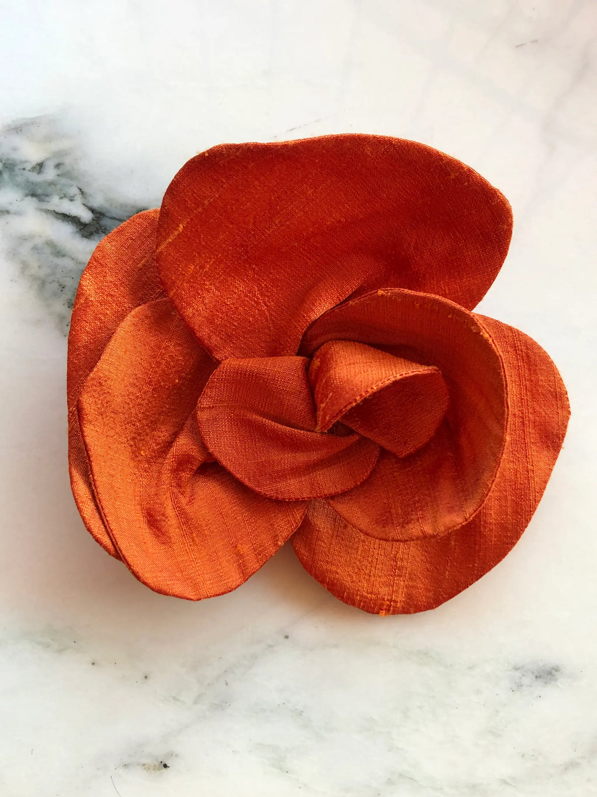 Broochella Bellucci small orange silk flower brooch with sculptural layered petals and rich silk sheen, photographed on a white marble background with grey veining.