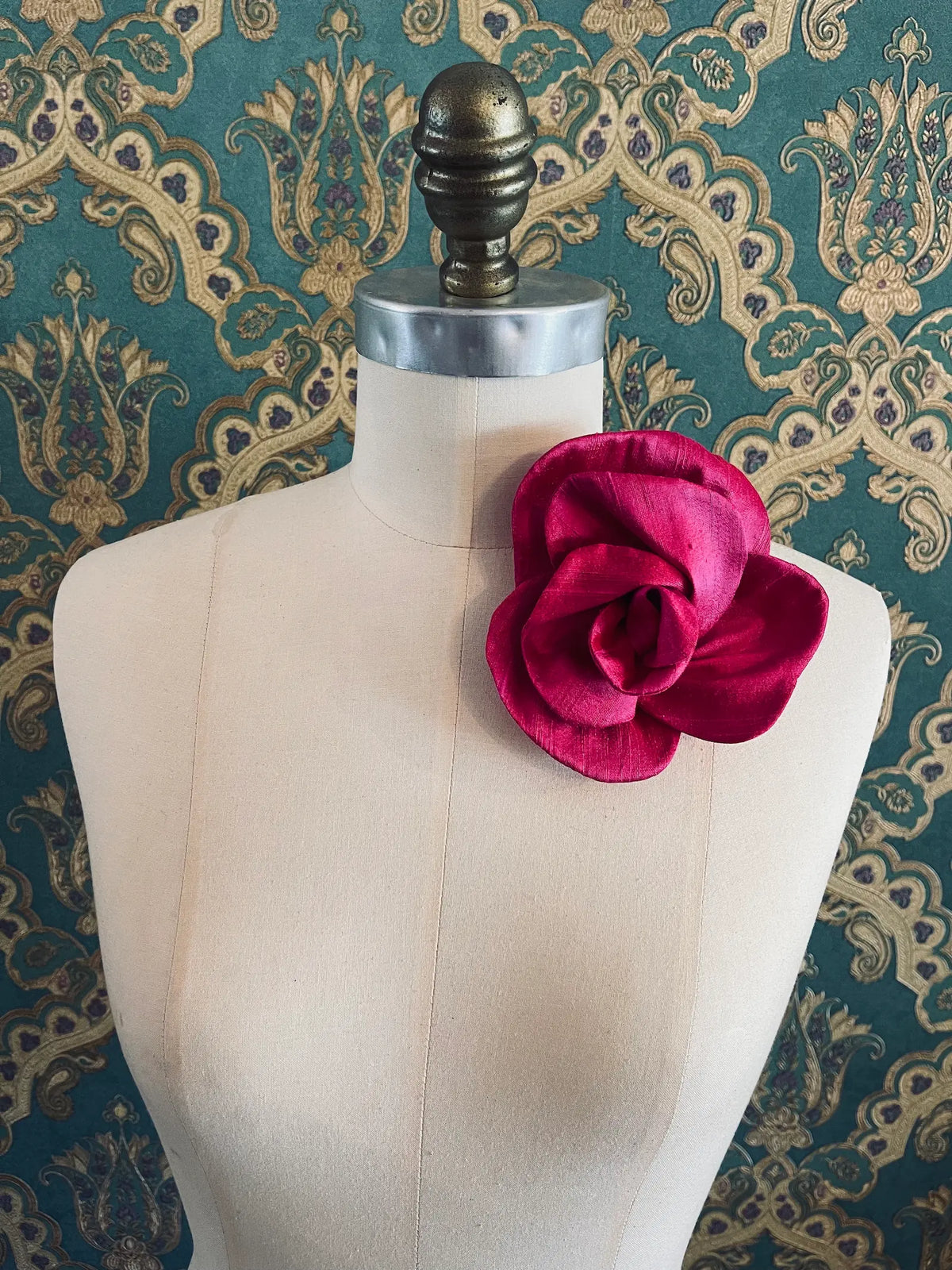 Broochella Bellucci small bright pink silk flower brooch with sculptural layered petals, styled on a cream mannequin against ornate teal wallpaper.