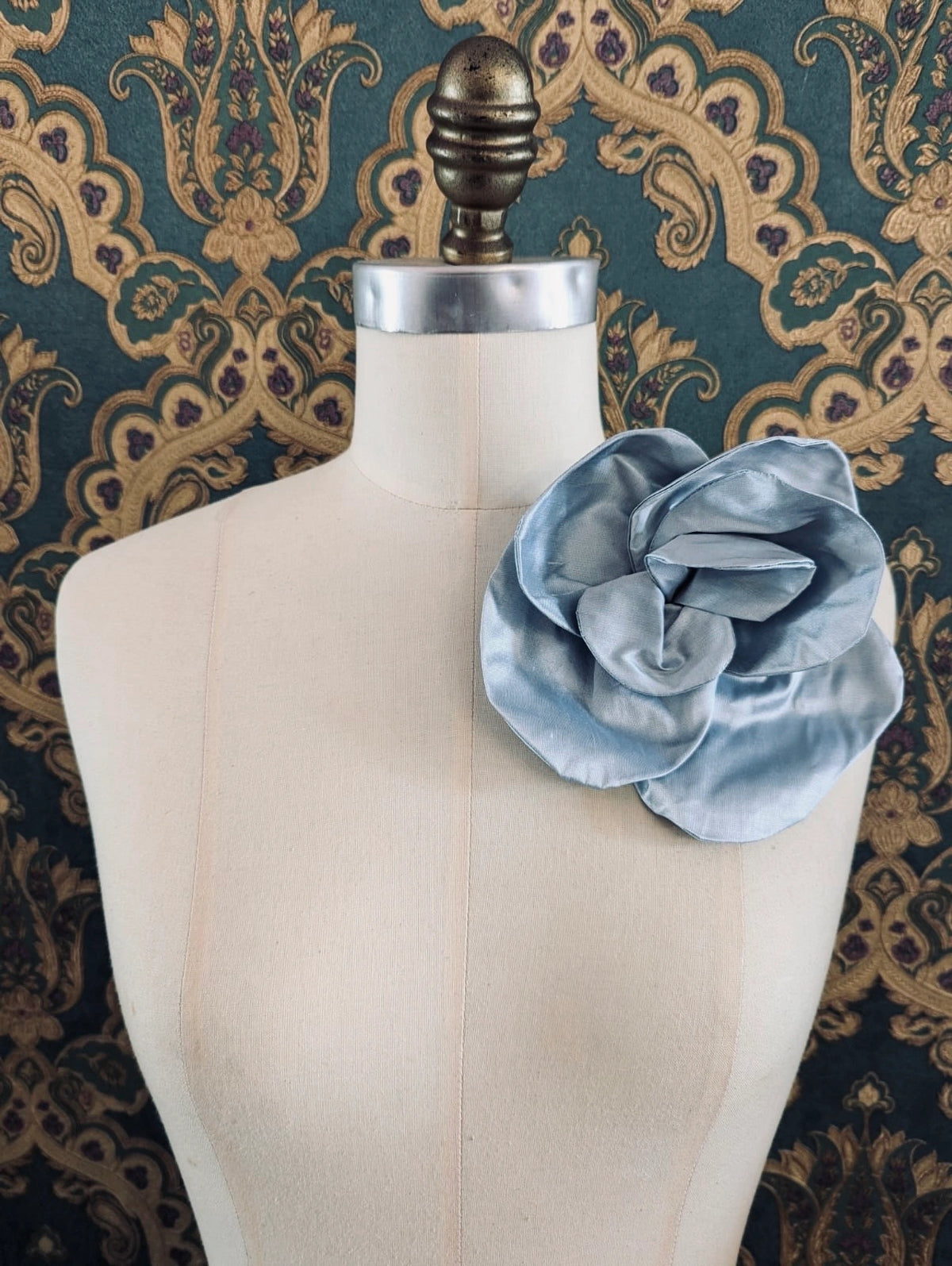 Broochella Bellucci small powder blue silk flower brooch with softly sculpted petals, styled on the shoulder of a cream mannequin against ornate teal wallpaper.