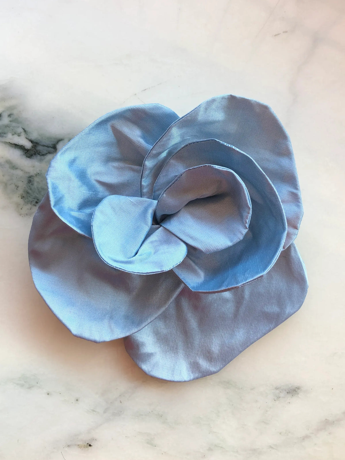 Broochella Bellucci small powder blue silk flower brooch with softly curved sculptural petals, photographed on a white marble background with grey veining.