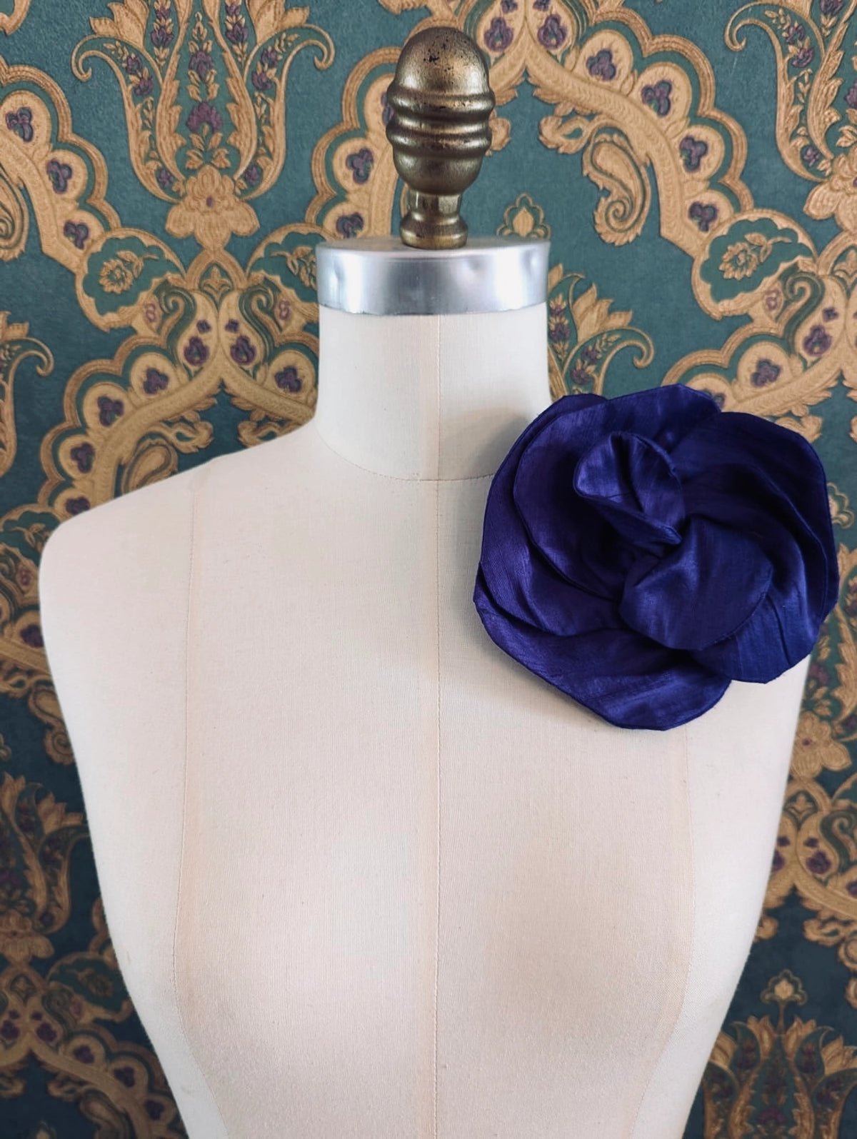 Broochella Bellucci small purple silk flower brooch with dramatic layered petals, styled on a cream mannequin against an ornate teal wallpaper background.