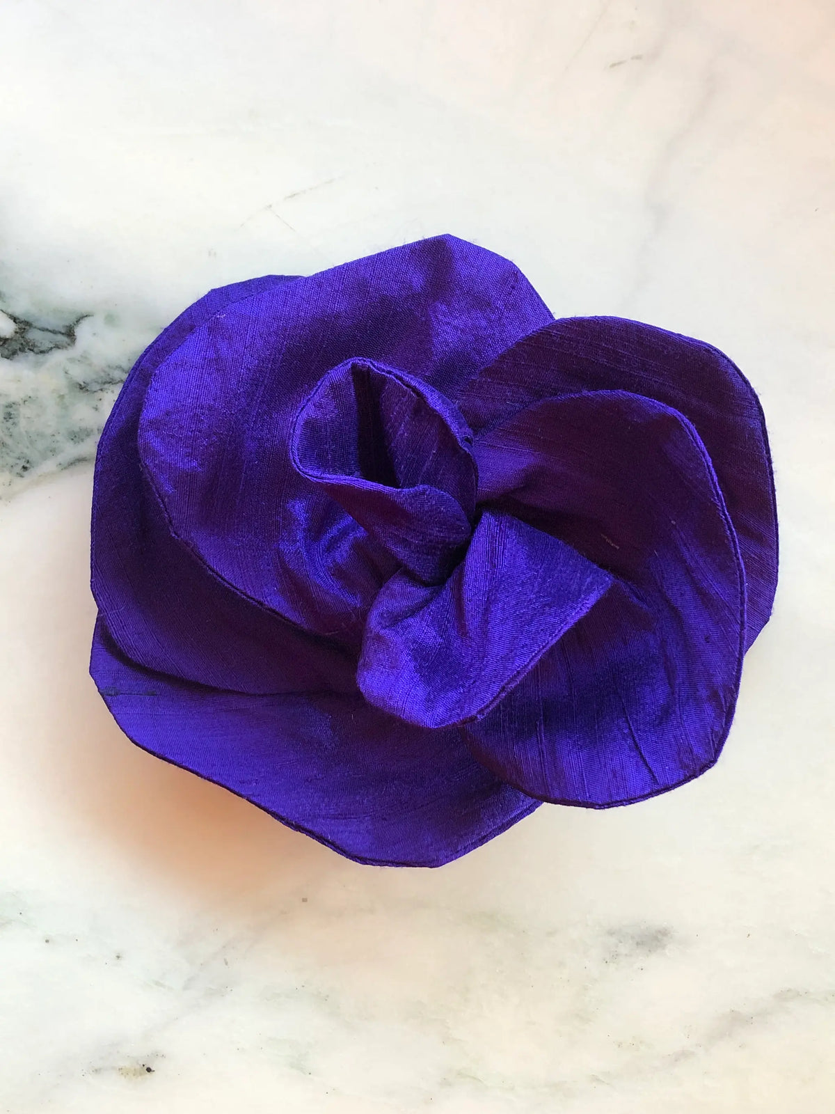 Broochella Bellucci small purple silk flower brooch with dramatic sculptural petals and rich silk sheen, photographed on a white marble background with grey veining.