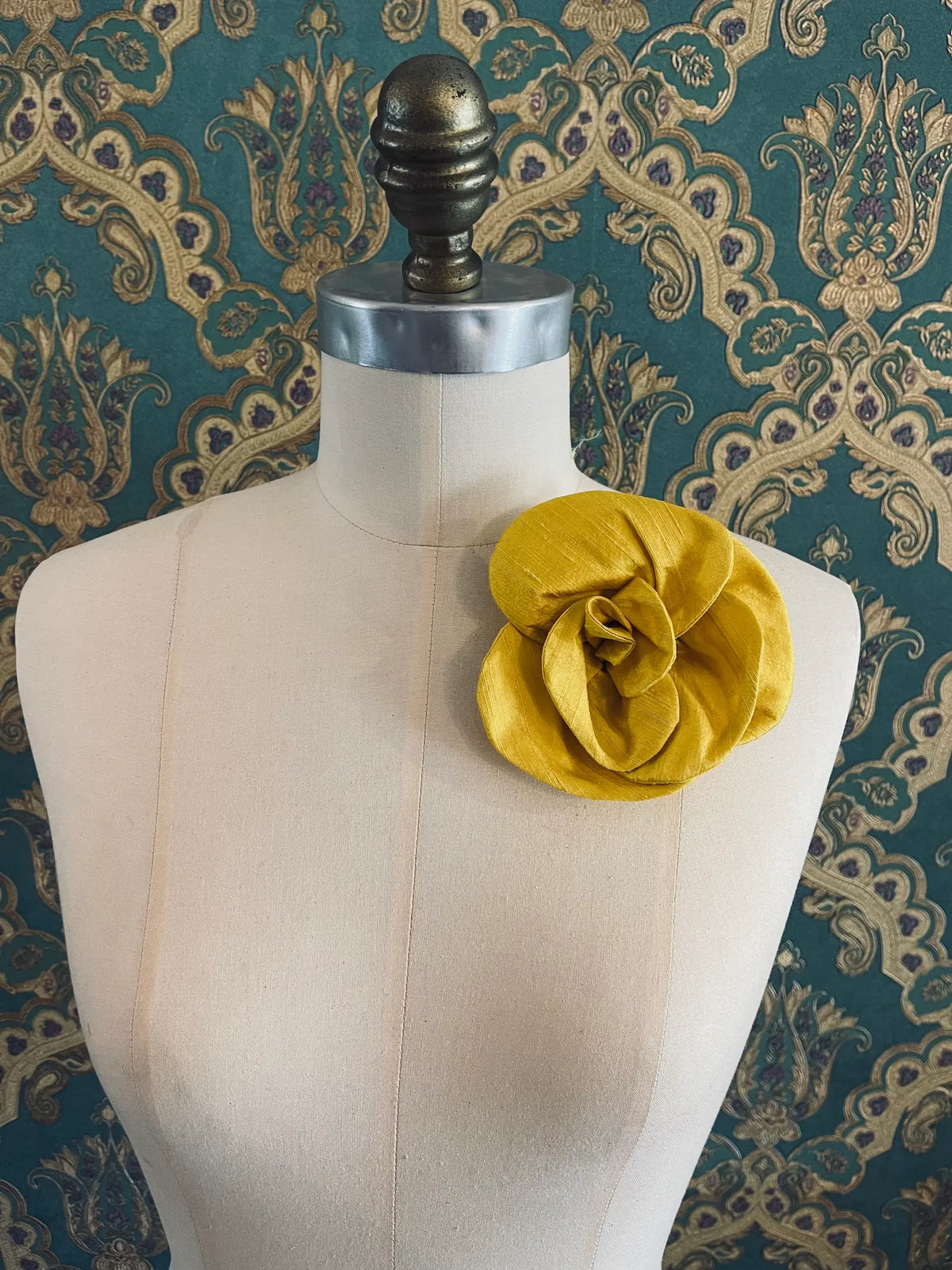 Broochella Bellucci small yellow silk flower brooch with lustrous sculptural petals, styled on the shoulder of a cream mannequin against ornate teal wallpaper.