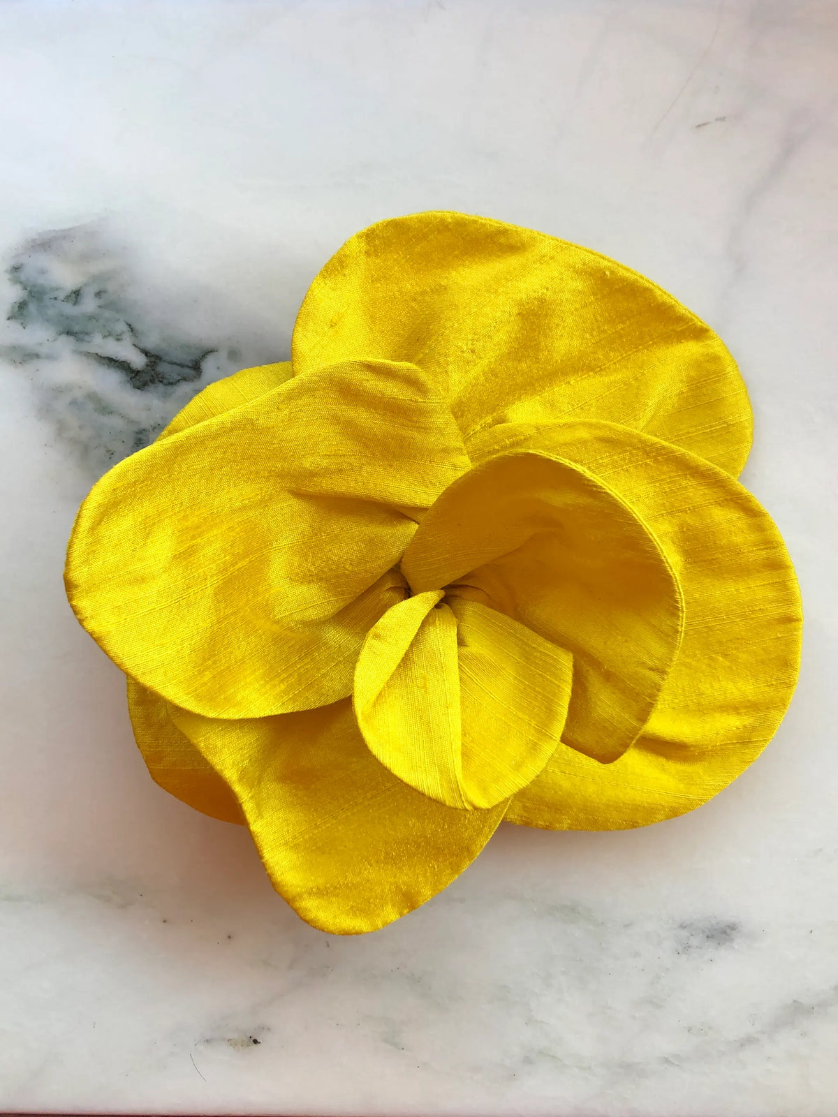 Broochella Bellucci small yellow silk flower brooch with bold layered petals and lustrous texture, photographed on a white marble background with grey veining.