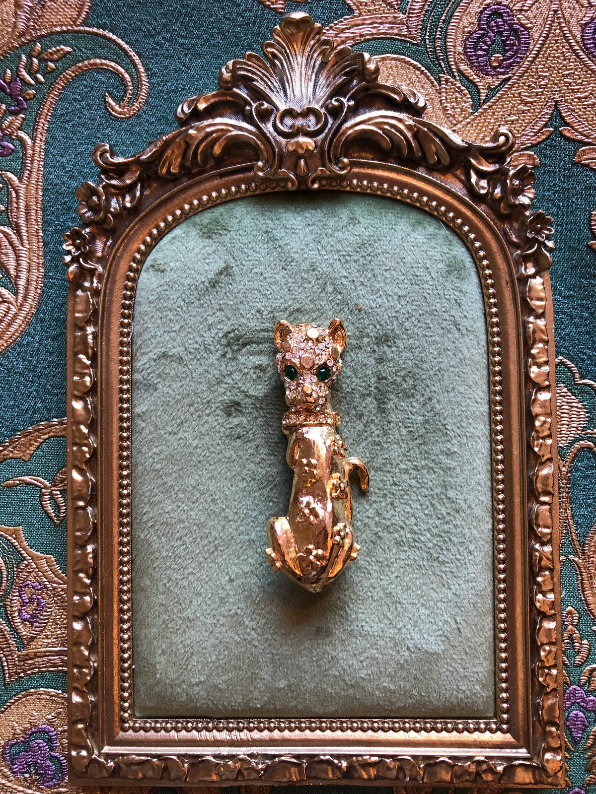 Broochella Bianca Vintage Gold Cat Brooch featuring an elongated gold-tone cat with emerald-green eyes, clear crystal detailing and floral accents. It is presented on a green velvet tray with an ornate frame. The background is green and gold damask wallpaper.