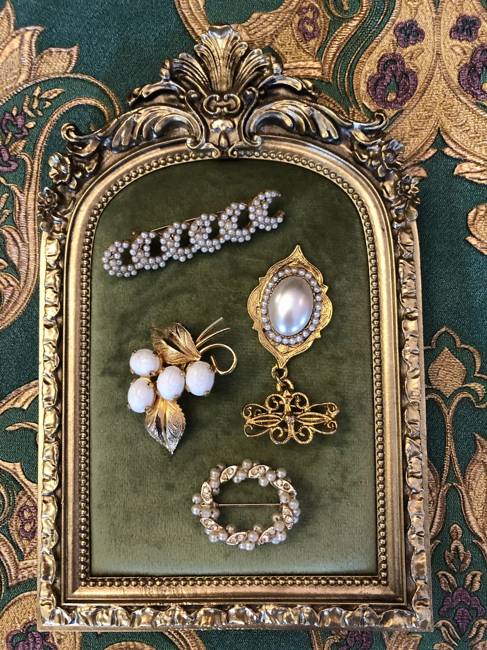 Broochella The Bianca Vintage Brooch Quartet featuring four ornate vintage pearl, ivory and gold-tone brooches with botanical, scroll and filigree details. It is presented on a green velvet tray with an ornate gold frame. The background is green and gold damask wallpaper