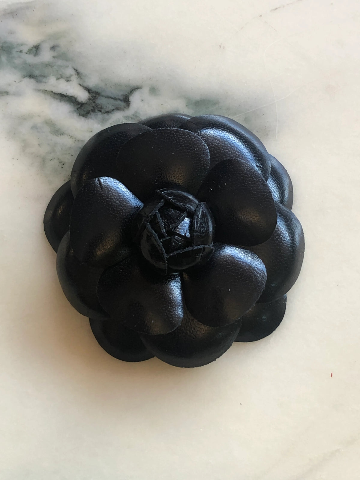 A Broochella Camelia handmade black leather flower-shaped accessory brooch,  resting on a white marble surface.