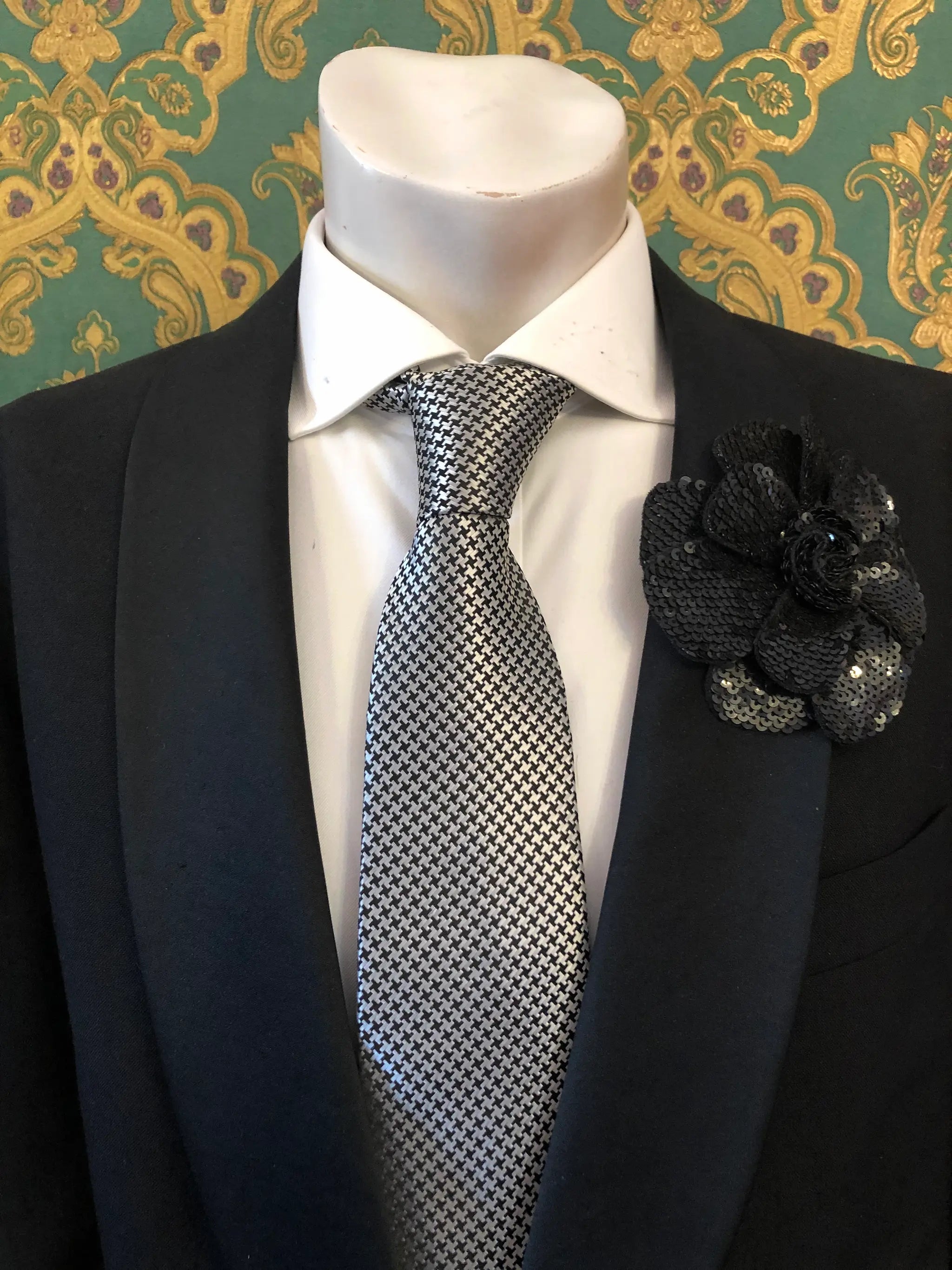 A men's formal accessory set displayed on a white mannequin torso, featuring a black suit jacket, a white dress shirt, a silver and black checkered tie, and an orange Men's Broochella Scintillante sequinned flower brooch lapel pin against a green and gold patterned wallpaper background.